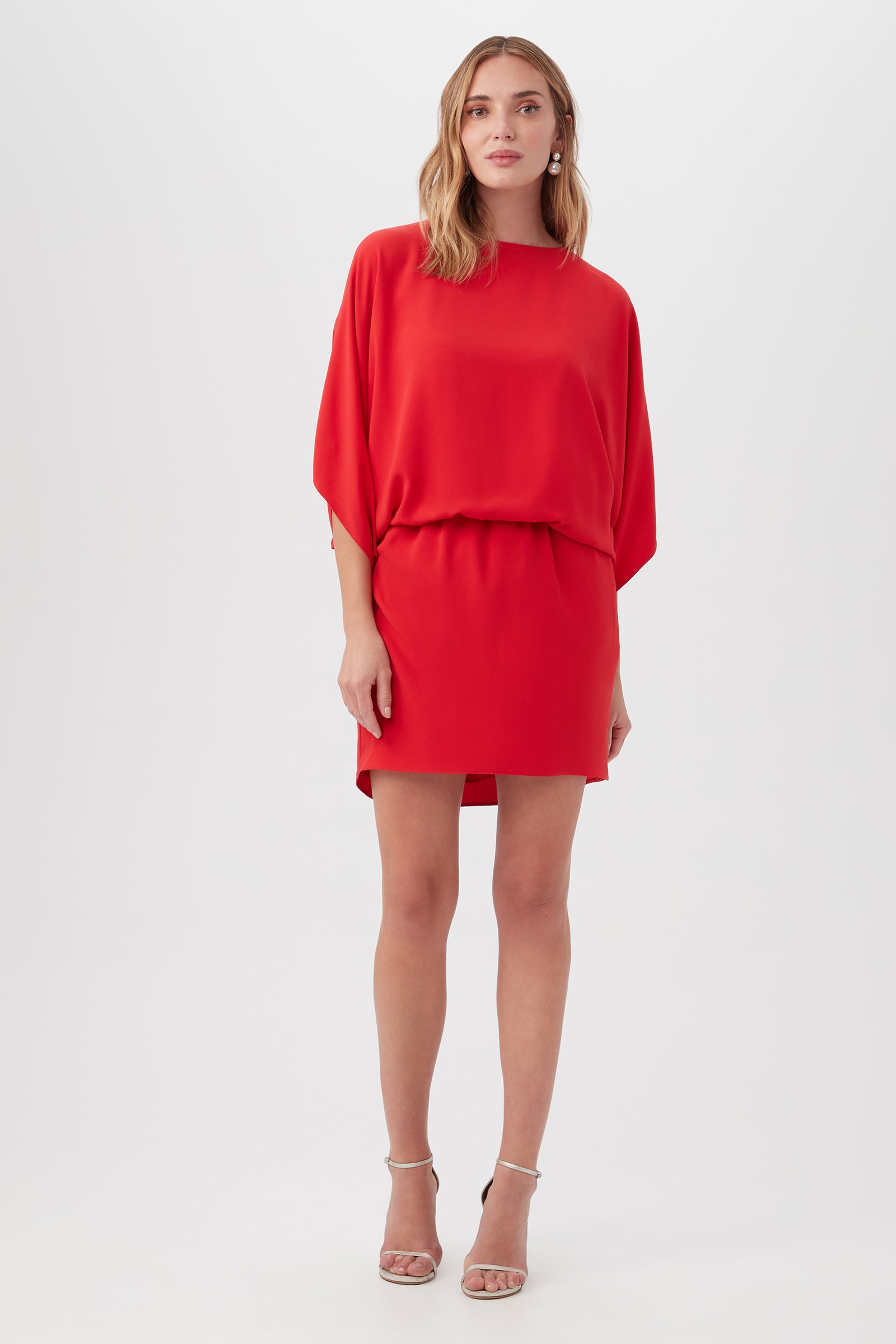 A woman faces forward, wearing the Manhattan Dressâ€”a loose-fitting, bright red short dress with elbow-length sleeves and a cinched waistâ€”and strappy beige high-heeled sandals against a plain white background.