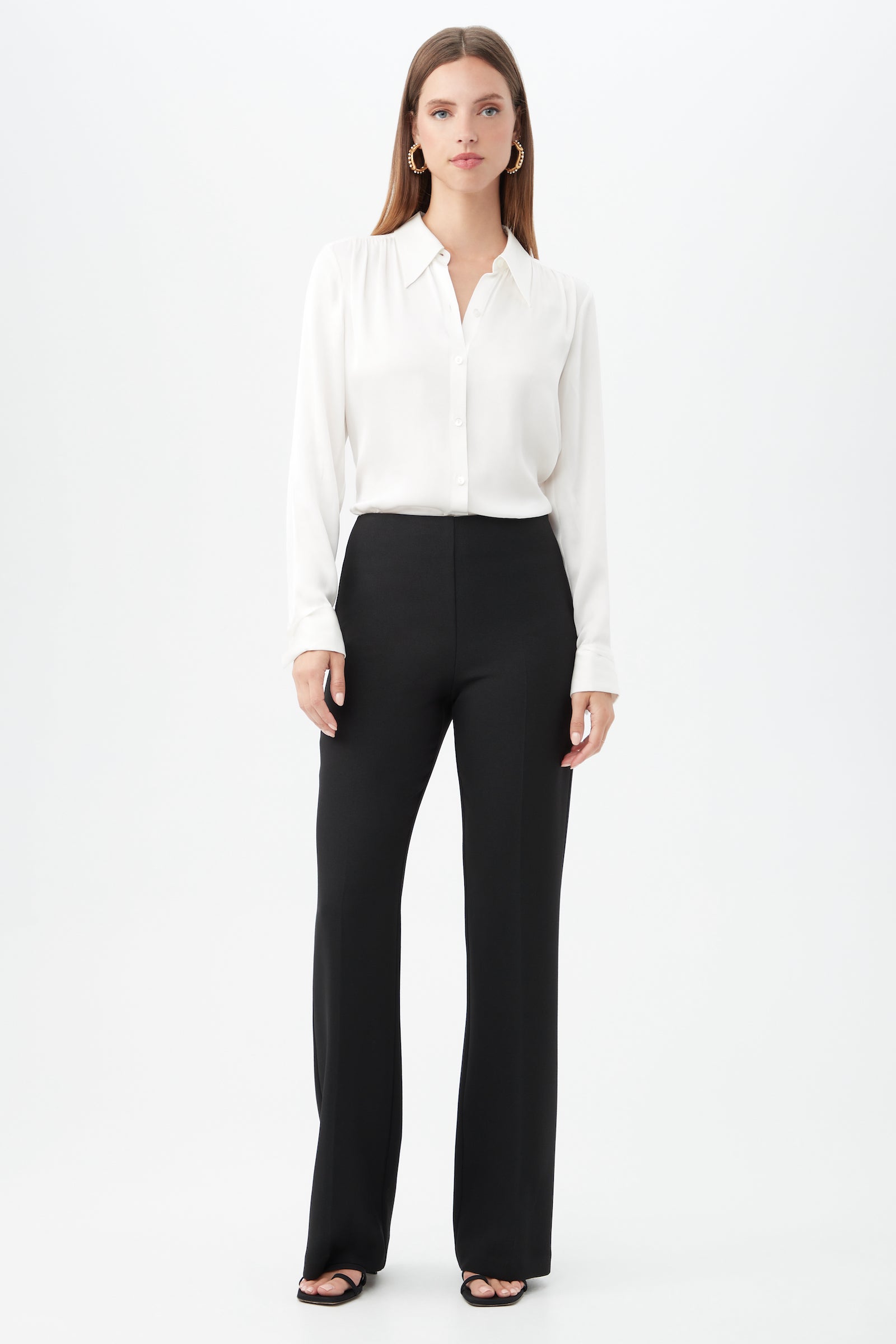 A woman stands against a plain white background wearing the Cassavetes Long-Sleeve Stretch Silk Button-Up Shirt, black high-waisted wide-leg pants, black open-toe heels, and gold hoop earrings. She has straight, shoulder-length hair.