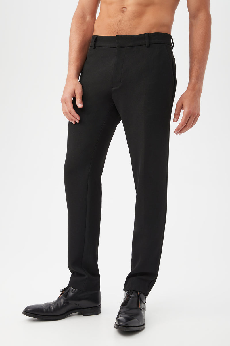 A man stands against a plain white background wearing the Clyde Slim Trouser - Hi-Line Suiting, made from sustainable fabric, paired with black dress shoes; his torso is bare and his arms rest relaxed at his sides.