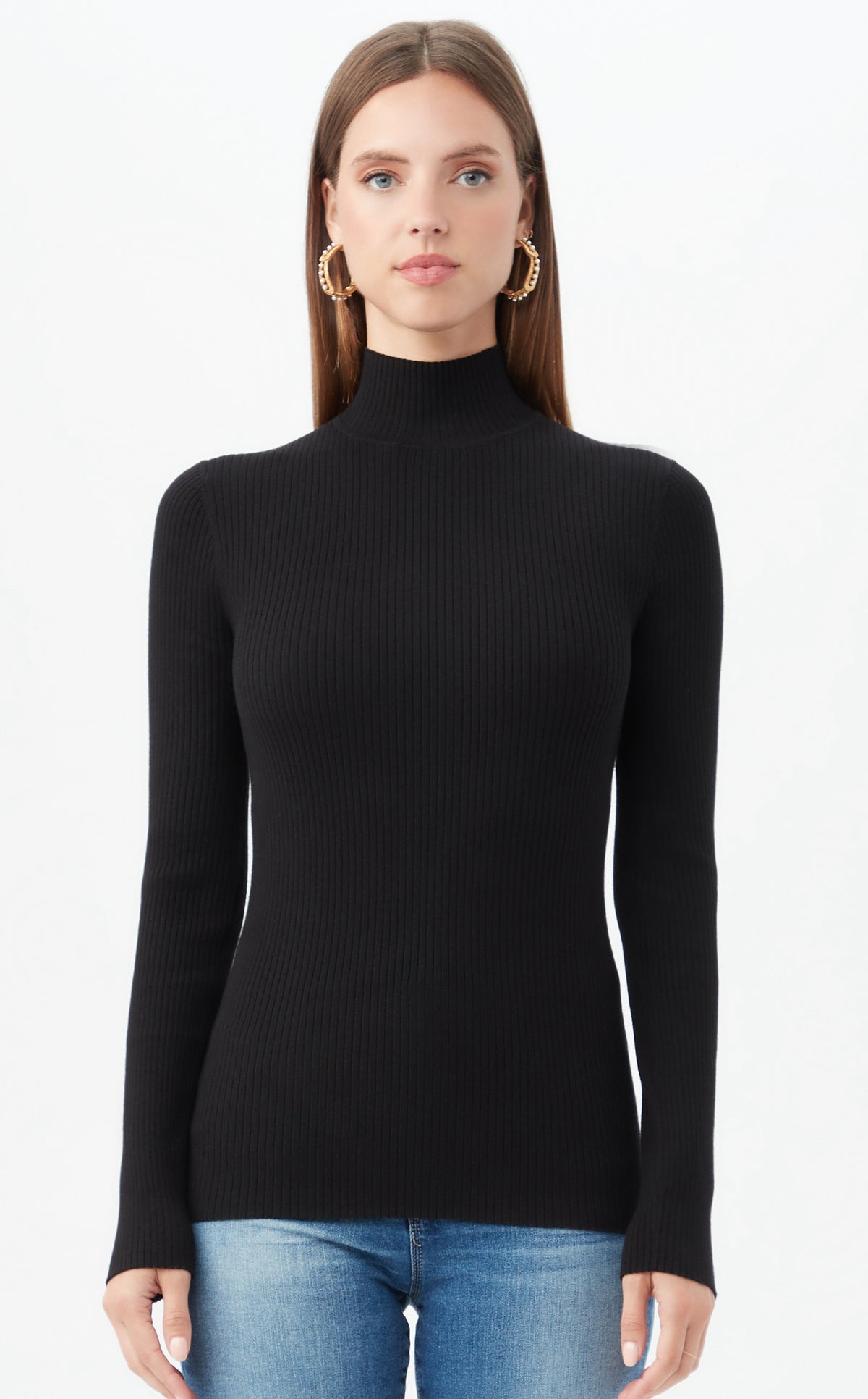 A woman with long brown hair, large gold hoops, and blue jeans models the black Hempstead Sweater—a fitted ribbed knit turtleneck—standing against a plain white background.