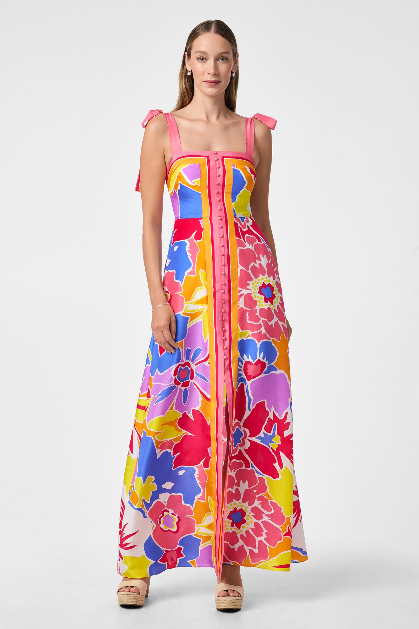 A woman with straight hair wears the Cami Sleeveless Dress - Soie Paradis, a long dress with shoulder ties and a vibrant floral pattern in pink, yellow, red, blue, and purple. She pairs it with platform sandals against a plain background.