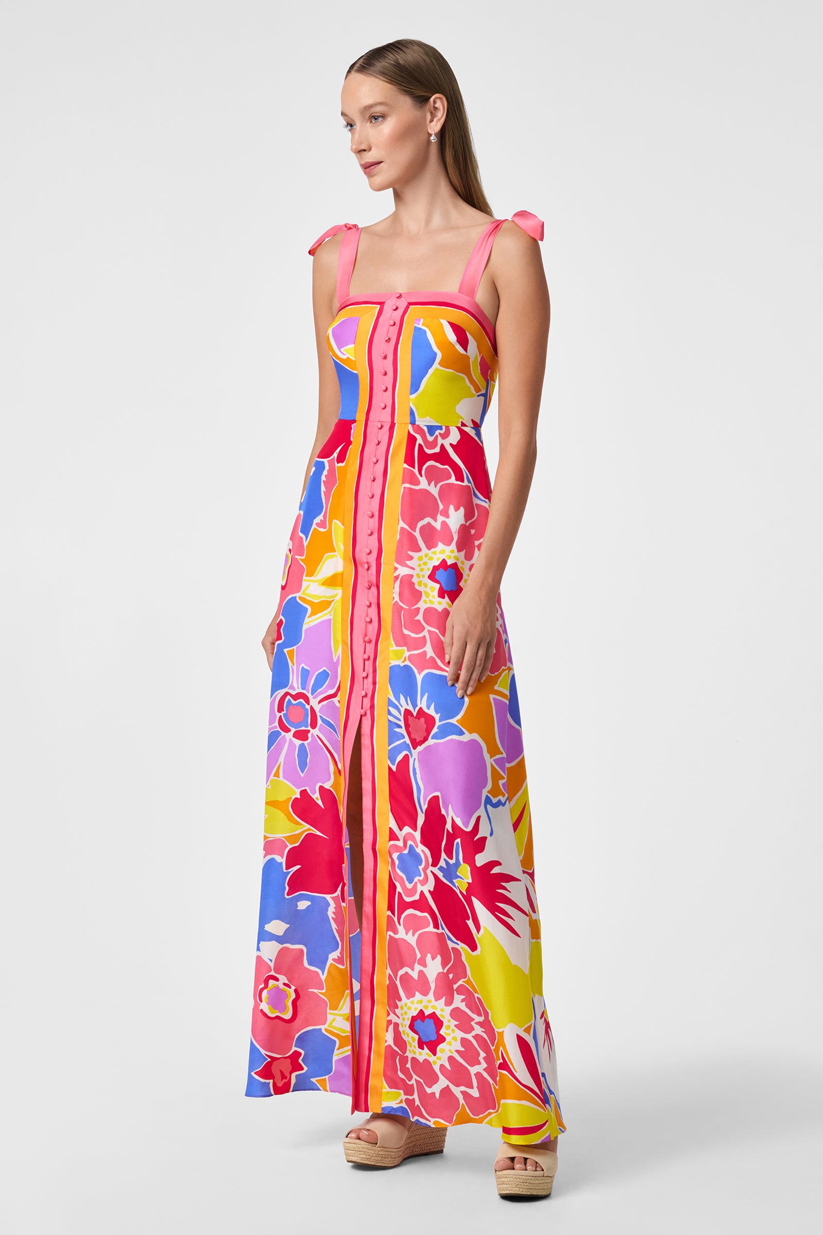 A woman wearing the Cami Sleeveless Dress - Soie Paradis, a colorful floral maxi with tie straps and front buttons, paired with wedge sandals, stands against a plain white background.