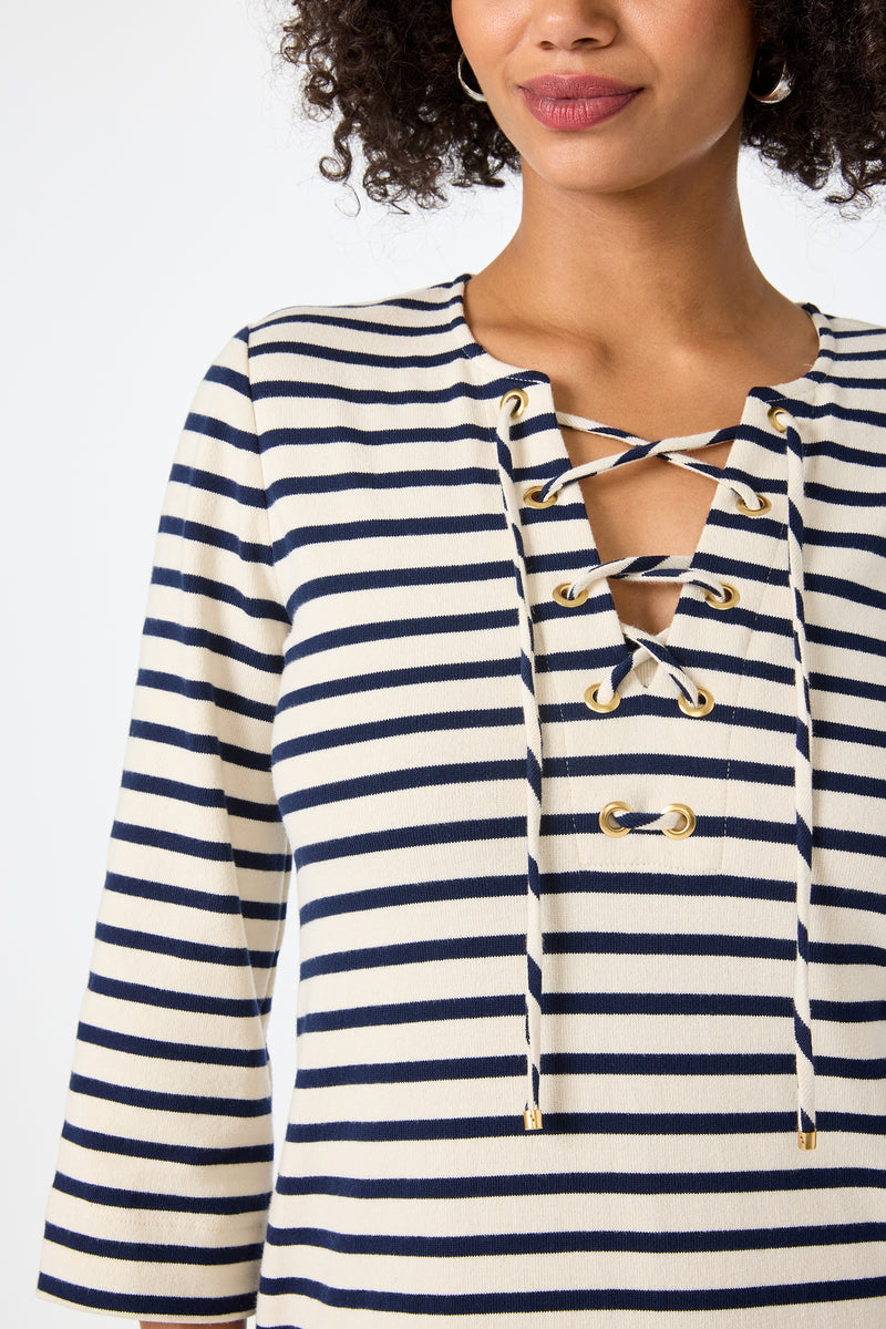 Woman wearing the Catamaran 3/4 Sleeve Maxi Dress in cream and navy blue stripes with a lace-up neckline and gold eyelets, shown from upper torso to mouth, smiling slightly.