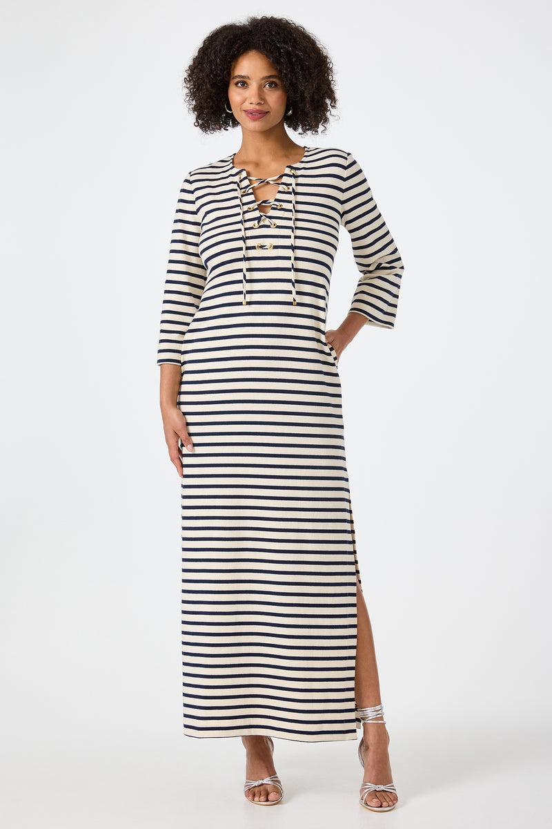 A woman wears the Catamaran 3/4 Sleeve Maxi Dress in Noh Nightfall Stripe with a lace-up neckline, one hand in her pocket and the other by her side, paired with silver sandals.