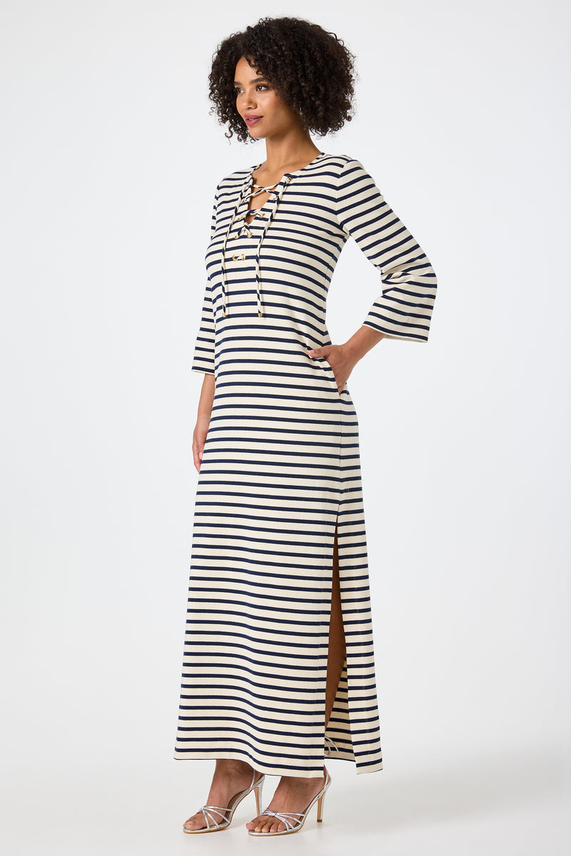A woman with curly hair stands in profile wearing the Catamaran 3/4 Sleeve Maxi Dress, featuring black and white stripes, a lace-up neckline, side slit, and paired with strappy heeled sandals.