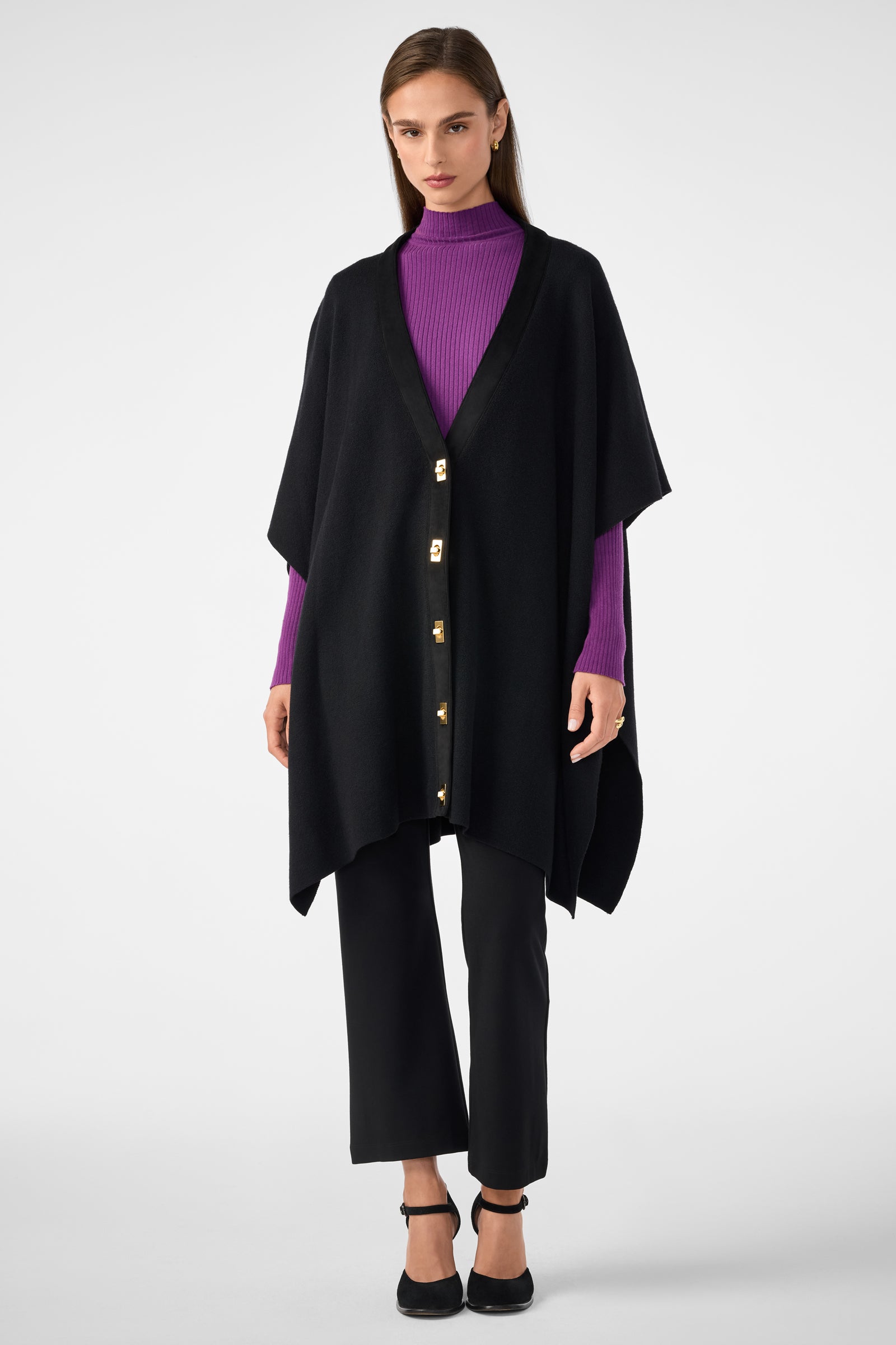 A woman with straight brown hair and a neutral expression stands against a plain background wearing the Sapphire Poncho, a loose black cape with gold buttons, over a purple ribbed turtleneck, paired with black pants and heels.