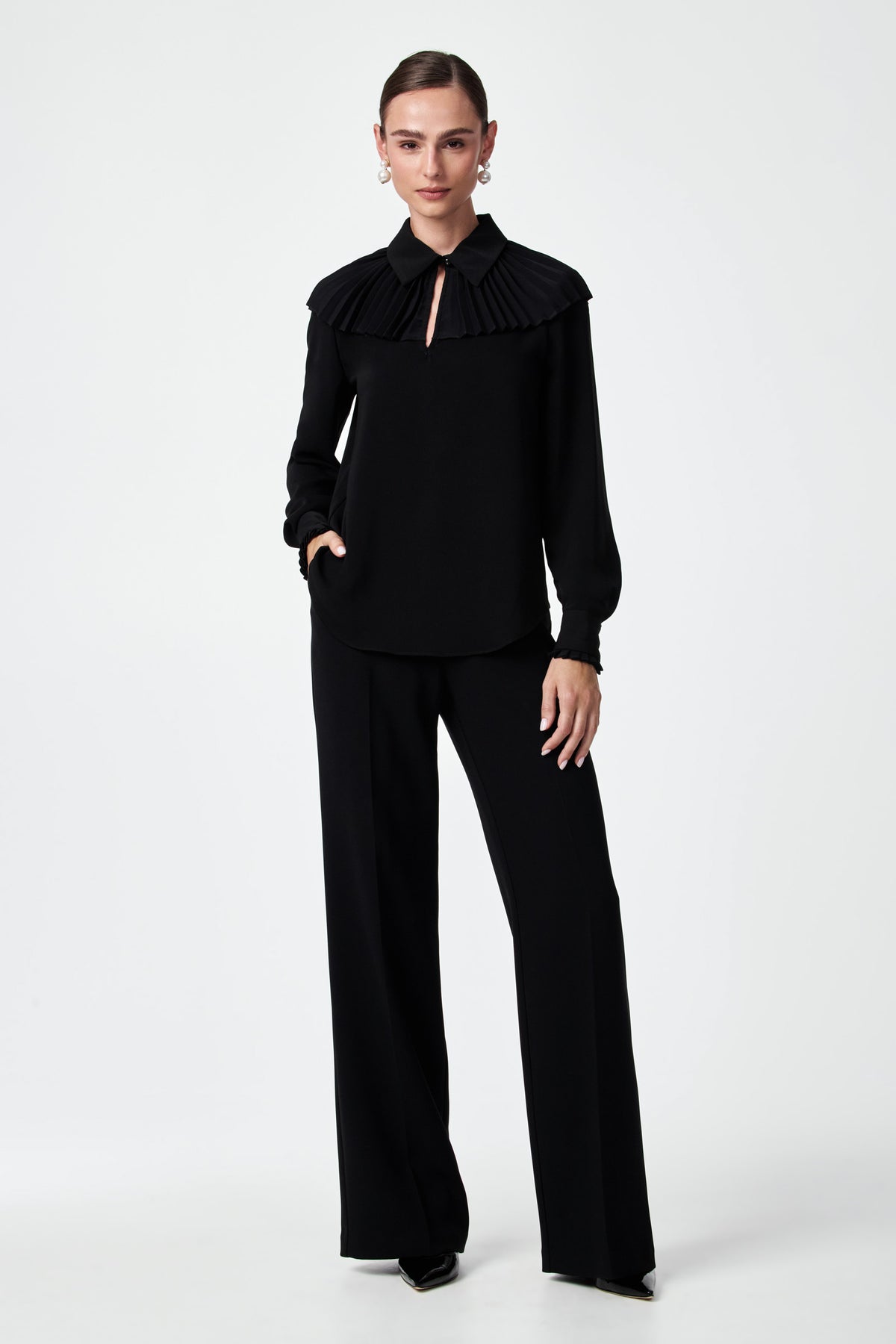 A woman stands against a plain white background wearing the black Windmill Top with pleated detailing and keyhole neckline, paired with wide-leg black trousers and black heels. She has one hand in her pocket.