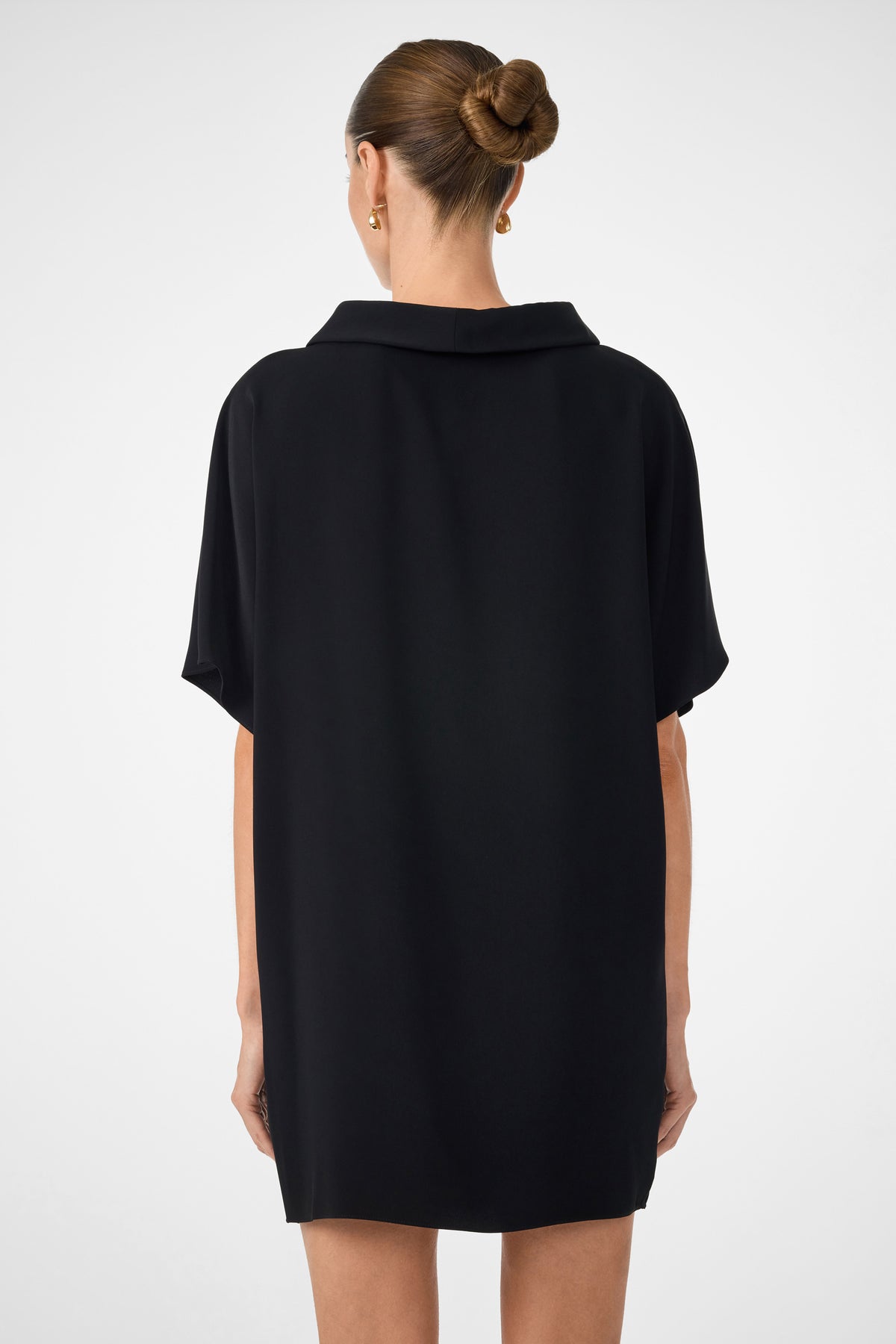 A woman with brown hair in a neat bun wears the black Honeysuckle Short Dolmain Sleeve Shift Dress, facing away from the camera against a plain light background.