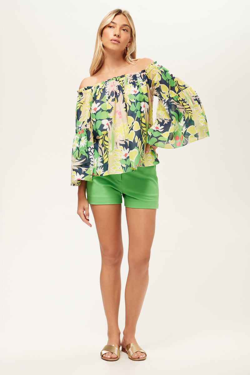 A woman wears the Perry Off-Shoulder Top - PS Eternal, featuring a colorful floral design with wide sleeves, paired with bright green shorts and tan sandals, standing against a plain light background.