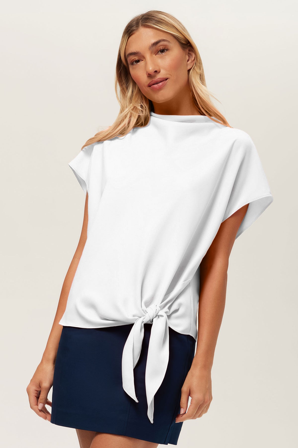 A woman wearing the Bisque Top, a white short-sleeve blouse with a side waist tie, paired with a navy blue skirt, stands against a plain light background.