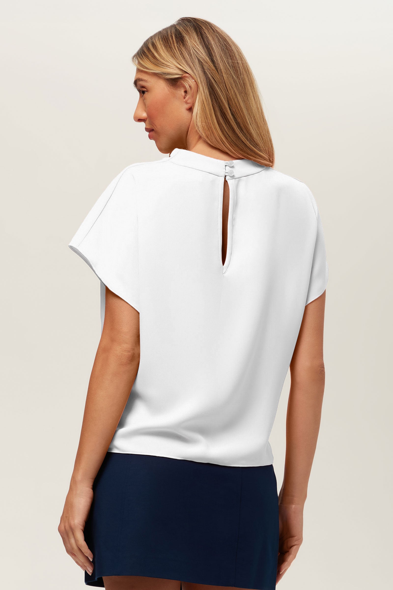 A woman with long, straight blonde hair is seen from the back wearing the Bisque Top—a white short-sleeve blouse featuring a keyhole button neckline—paired with a navy skirt against a plain light background.