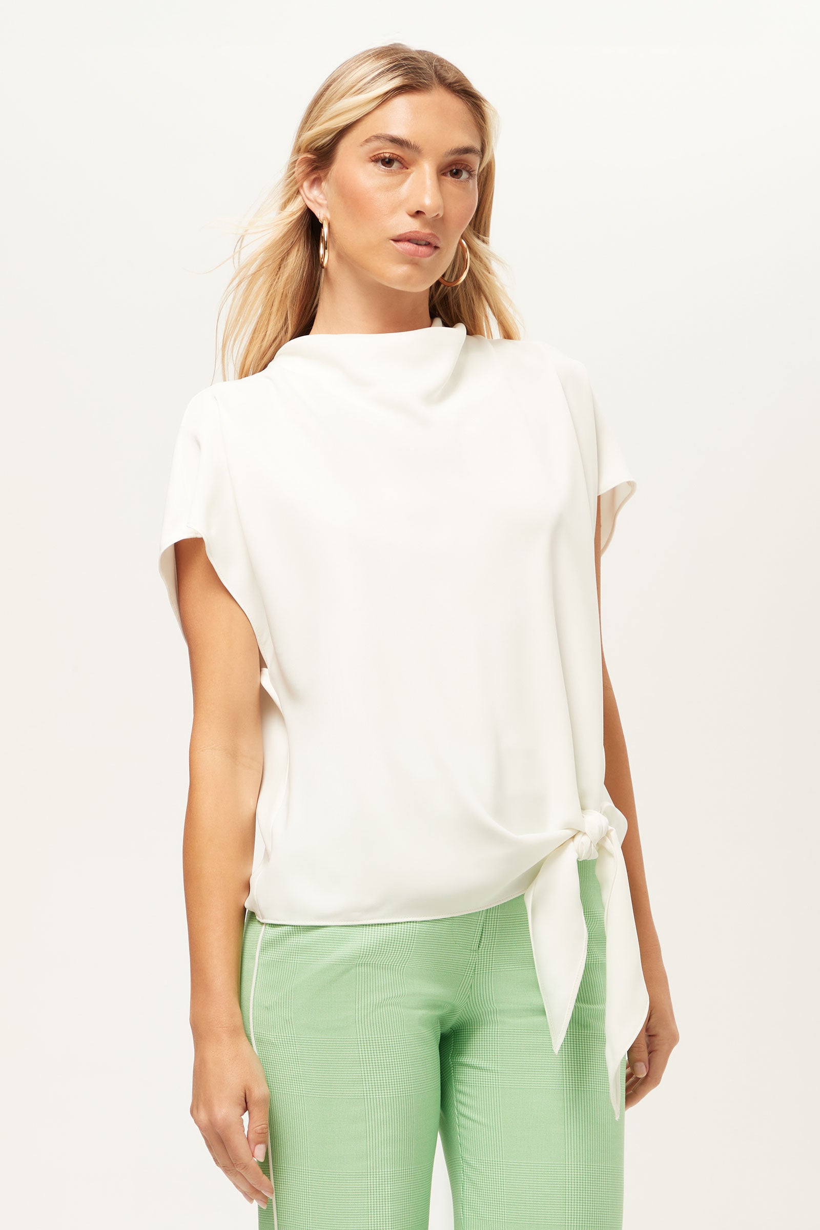 A woman with long blonde hair wears the Bisque Short-Sleeve Funnel Neck Top in Whitewash, paired with light green pants, standing against a plain white background.