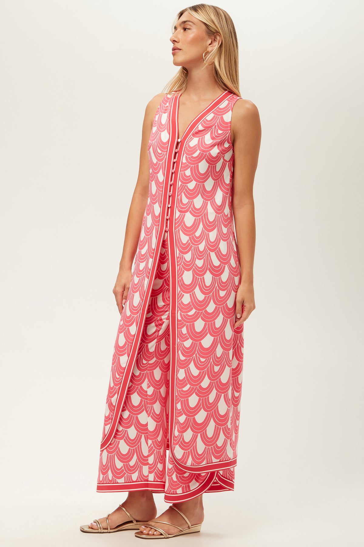 A woman wears the Slice Sleeveless Maxi Top - Ace Arches, an ankle-length pink and white dress with scalloped patterns, front buttons, and side slits. She has straight blonde hair and flat sandals.