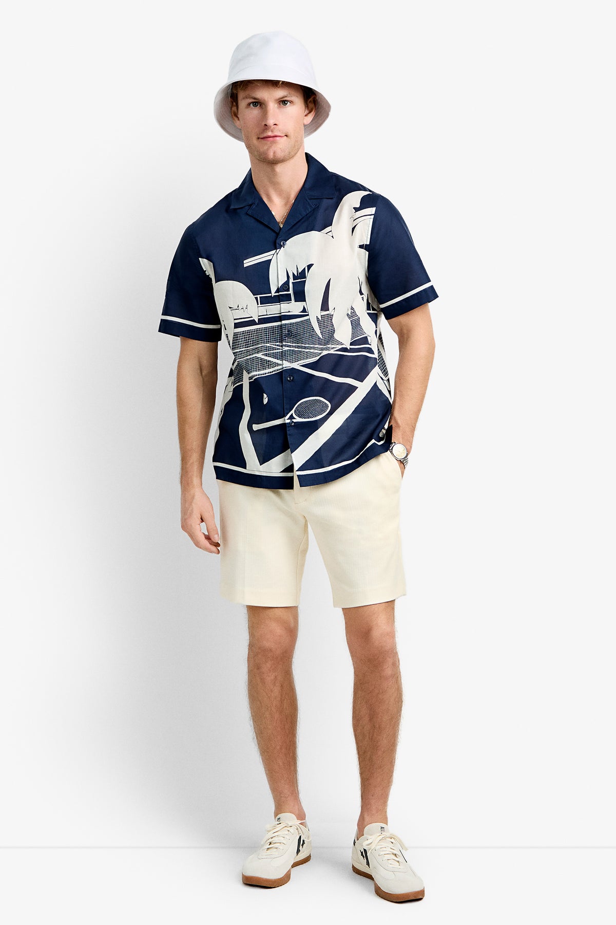 A man wearing the Josue Short-Sleeve Shirt - Tennis Print, cream shorts, a white bucket hat, white socks, and white sneakers stands against a plain white background.