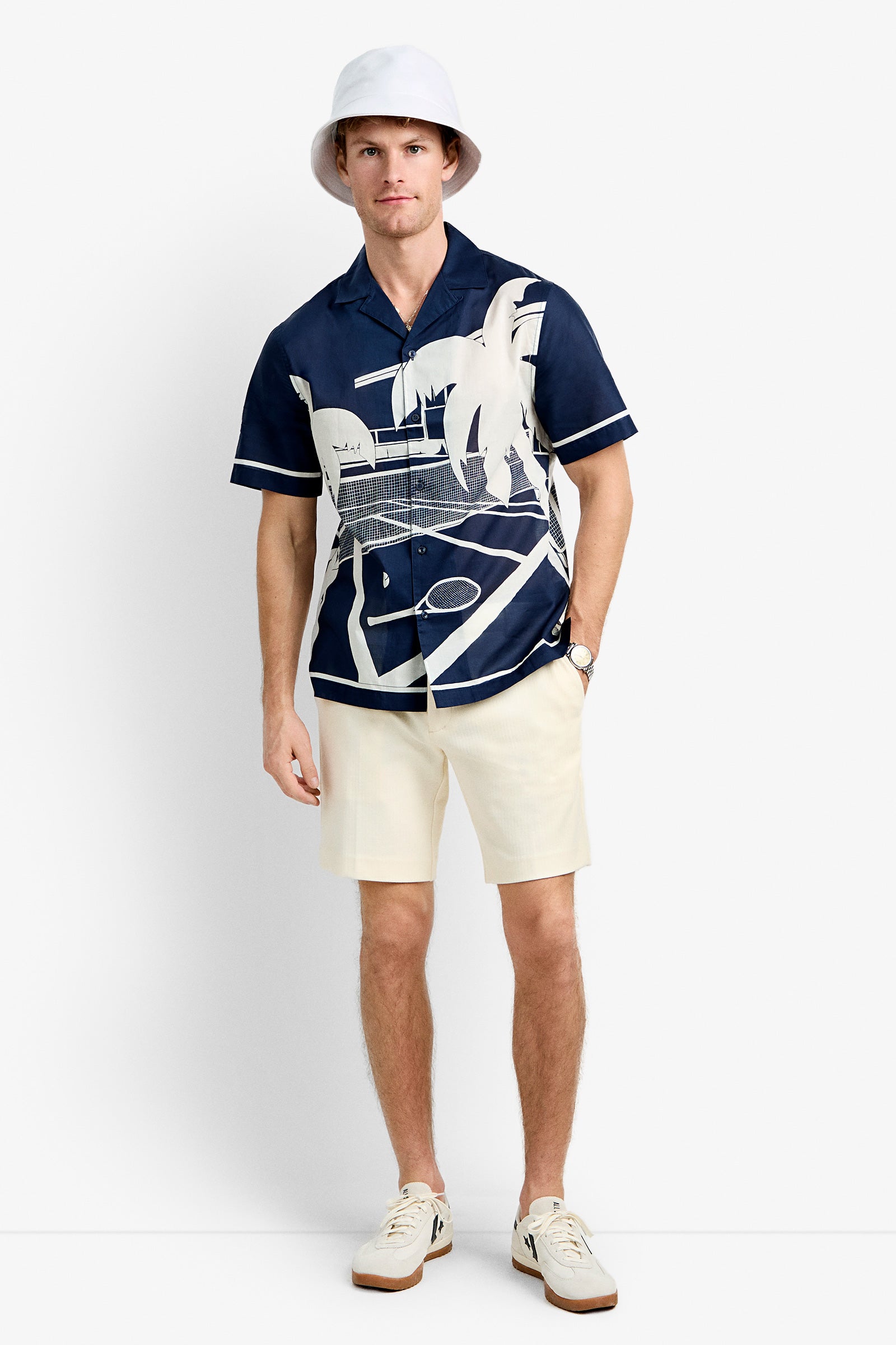 A man wearing the Josue Short-Sleeve Shirt - Tennis Print, cream shorts, a white bucket hat, white socks, and white sneakers stands against a plain white background.