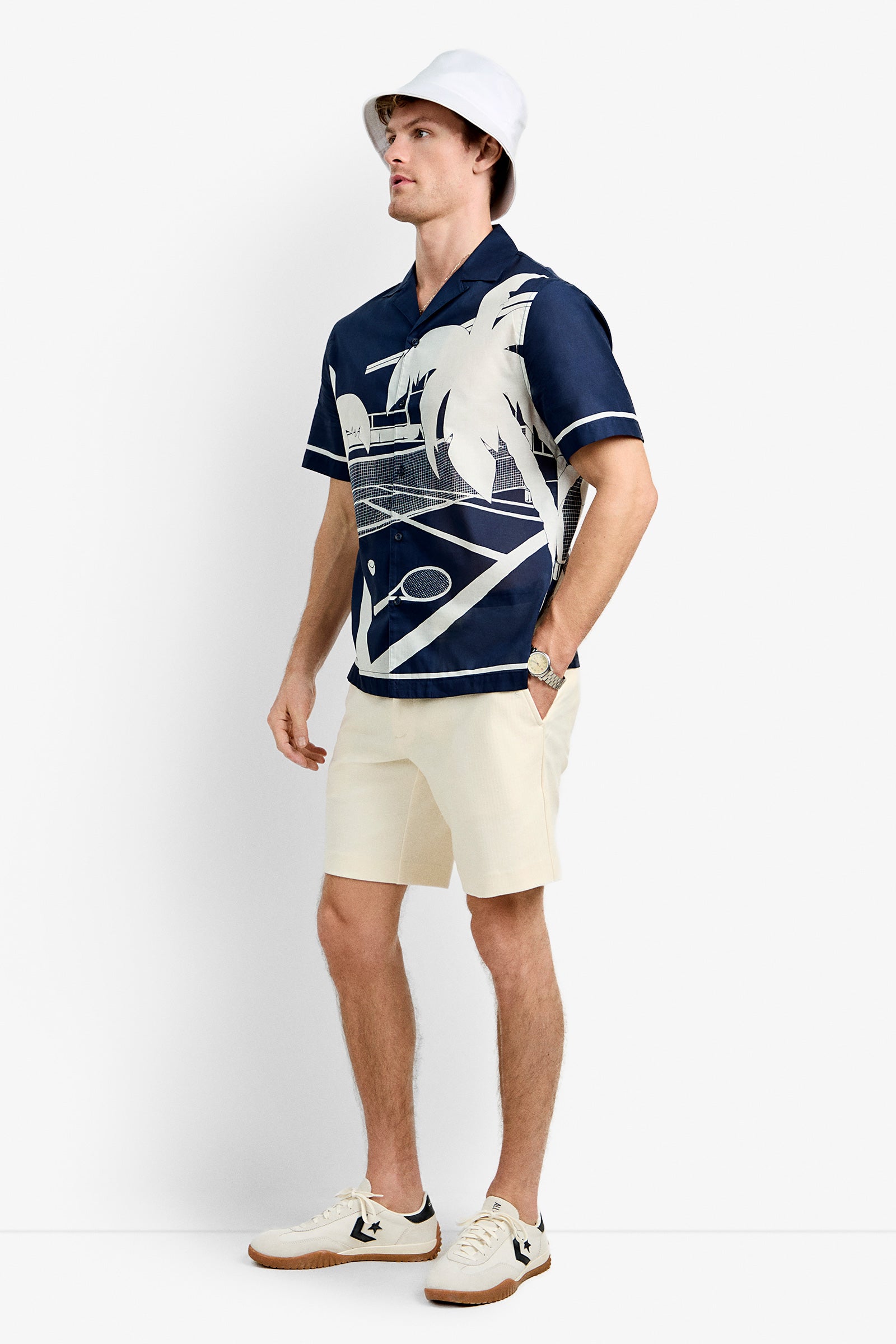 A man in a white bucket hat, Josue Short-Sleeve Shirt - Tennis Print (navy blue), beige shorts, white sneakers with black accents, and a watch stands against a plain white background.