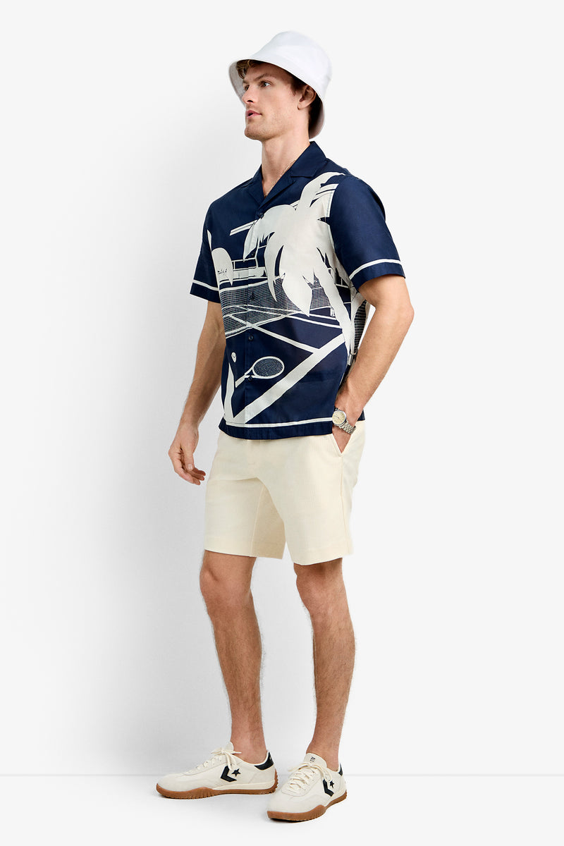 A man in a white bucket hat, Josue Short-Sleeve Shirt - Tennis Print (navy blue), beige shorts, white sneakers with black accents, and a watch stands against a plain white background.