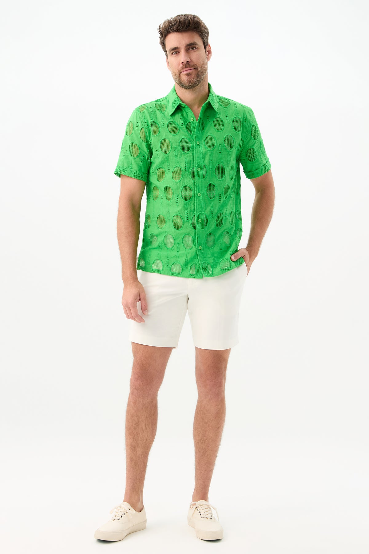 A man stands against a white background wearing the Branson Shirt - Tennis Eyelet, a bright green, short-sleeve, sheer button-up with large polka dots, paired with white shorts and sneakers. One hand is in his pocket and he looks relaxed.
