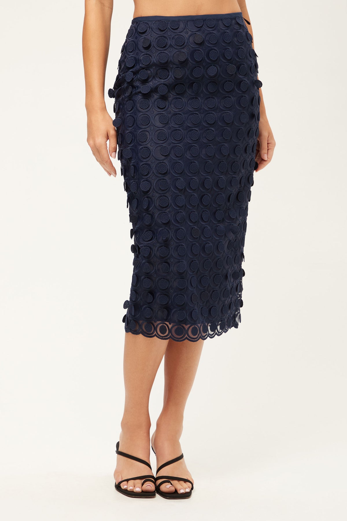 A woman wears the Dita 2 Midi Pencil Skirt - Agassi Embroidery in Ink, featuring a textured circular pattern. Paired with black strappy sandals, only her lower half is visible against a plain background.
