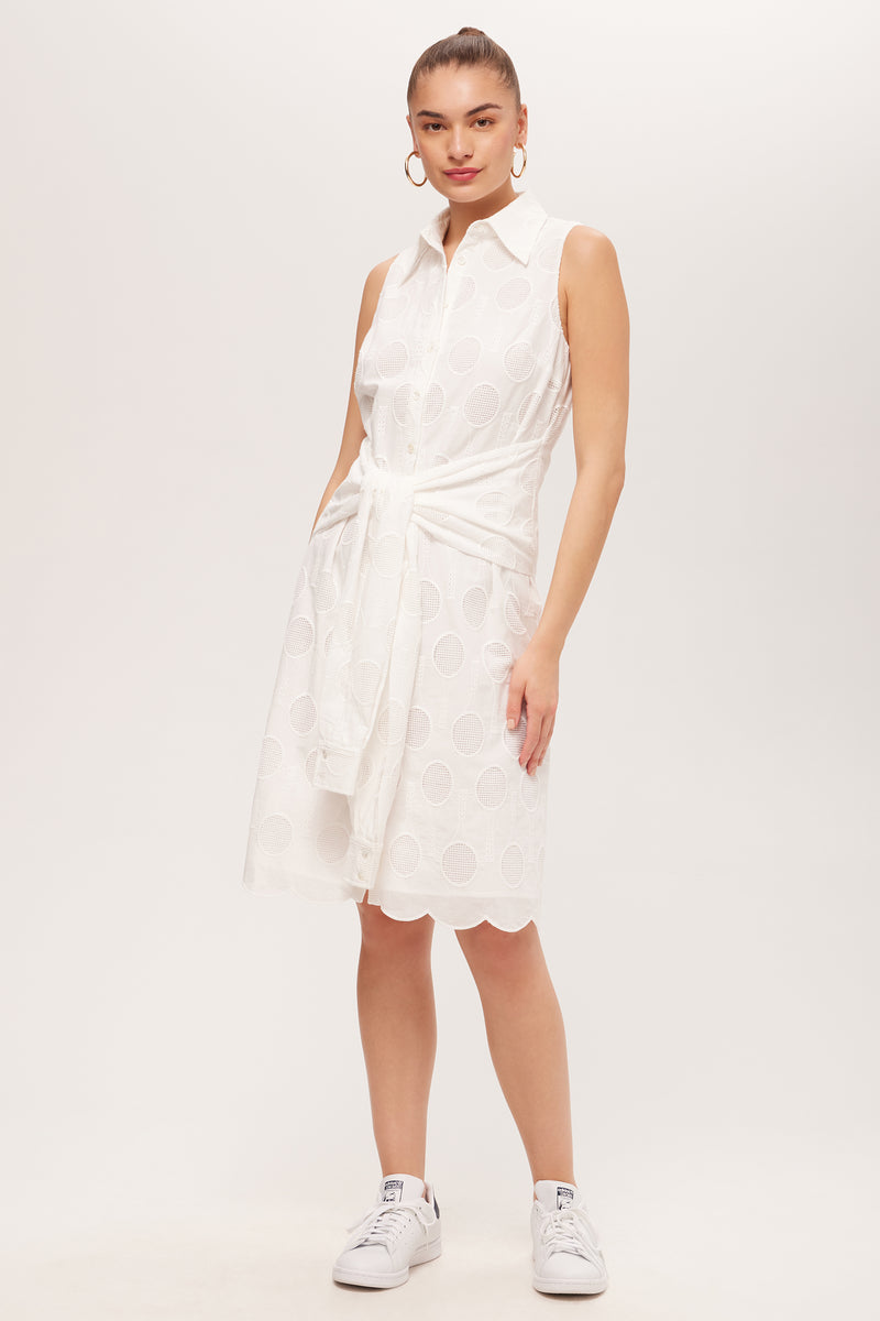 A woman stands against a plain background wearing the Rally Sleeveless Shirt Dress - Tennis Eyelet in Whitewash, featuring a tied waist, scalloped hem, and circular patterns. She pairs it with white sneakers and large hoop earrings.