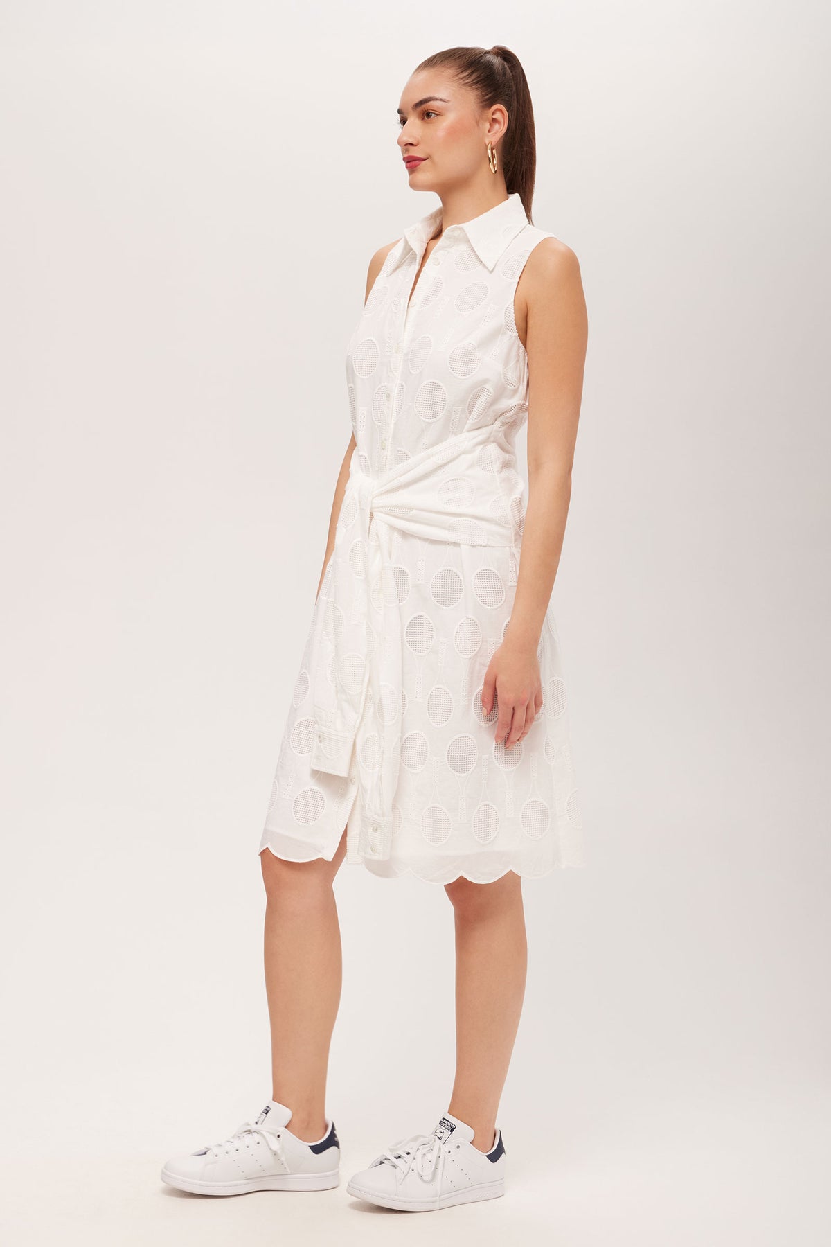 A woman wears the Rally Sleeveless Shirt Dress - Tennis Eyelet with white sneakers and a white sweater tied at her waist, posing against a plain light background.