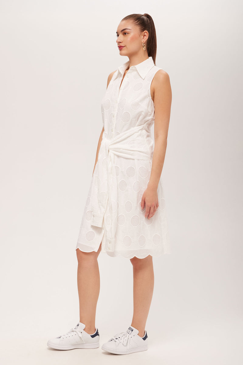 A woman wears the Rally Sleeveless Shirt Dress - Tennis Eyelet with white sneakers and a white sweater tied at her waist, posing against a plain light background.