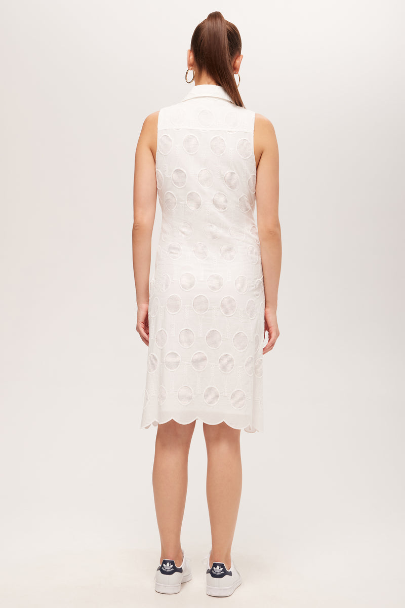 A woman with a high ponytail faces away, wearing the Rally Sleeveless Shirt Dress - Tennis Eyelet, featuring a white circular eyelet pattern and scalloped hem, paired with blue sneakers against a plain light background.