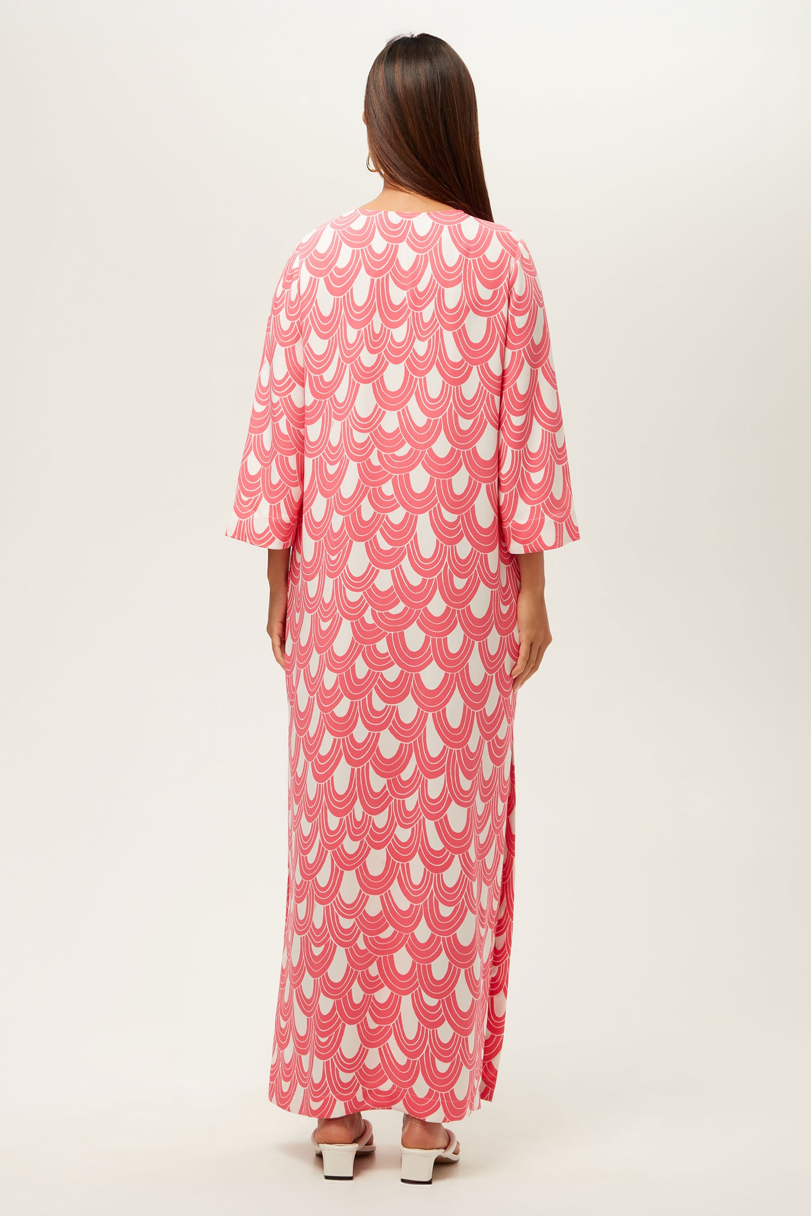 A woman stands facing away, wearing the Catamaran 3/4-Sleeve Maxi Dress featuring pink and white scallop patterns. She has straight brown hair, white sandals, and is set against a plain light background.