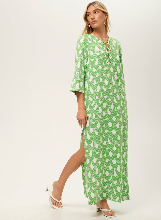 A woman in the Catamaran 3/4-Sleeve Maxi Dress with a green and white scallop pattern, lace-up neckline, and side slit stands against a plain background wearing white sandals, looking to the side.