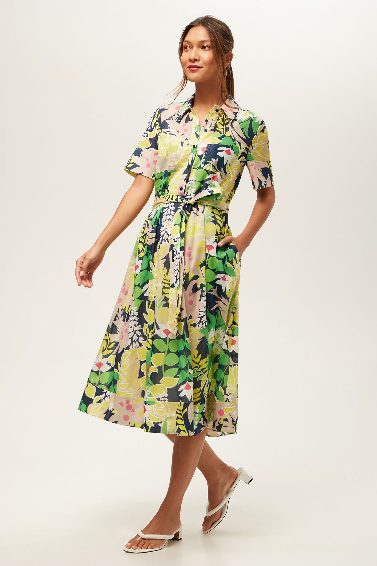 A woman wears the Misola 2 Short-Sleeve Midi Dress - PS Eternal, a colorful floral midi with a belted waist. She has long hair tied back, is smiling gently, and pairs the dress with white heeled sandals against a plain background.