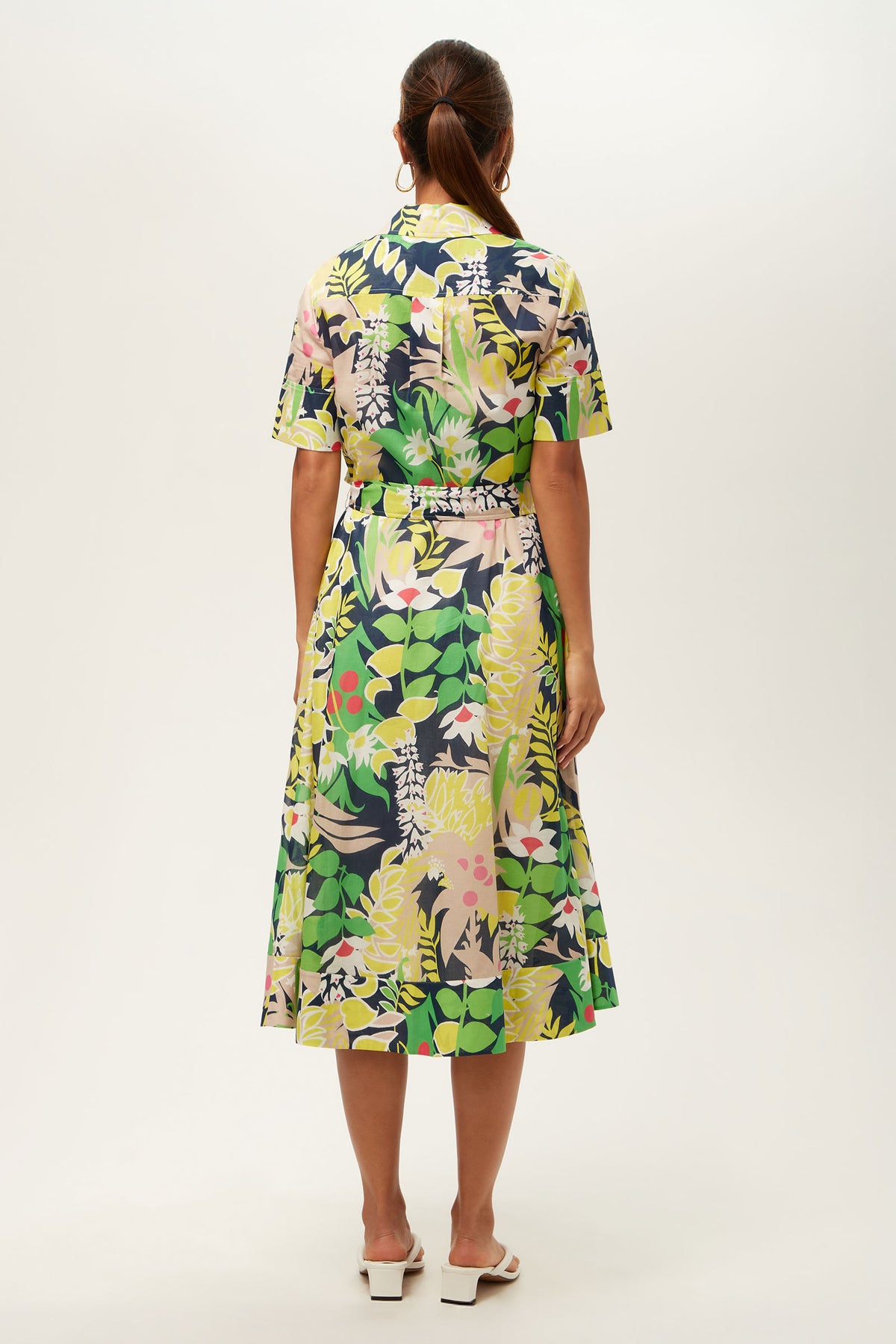 A woman with brown hair tied back wears the Misola 2 Short-Sleeve Midi Dress - PS Eternal, featuring a colorful bold floral and leaf print in green, yellow, pink, and navy. She pairs it with white heeled sandals.