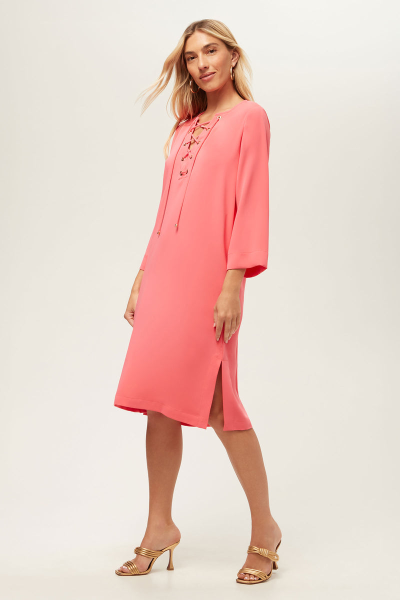 A woman poses against a plain light background wearing the Catamaran 3/4-Sleeve Midi Dress in Coral Crush, featuring a loose fit, lace-up front, side slit, and paired with gold strappy heeled sandals.