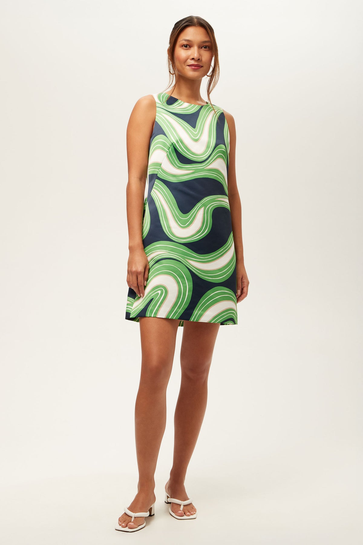 A woman stands against a plain background, wearing the Fairy Tale 2 Sleeveless Dress in Serena Satin—a knee-length dress with a bold green, navy, and white swirling pattern. Her long hair is tied back, and she pairs the look with white sandals.