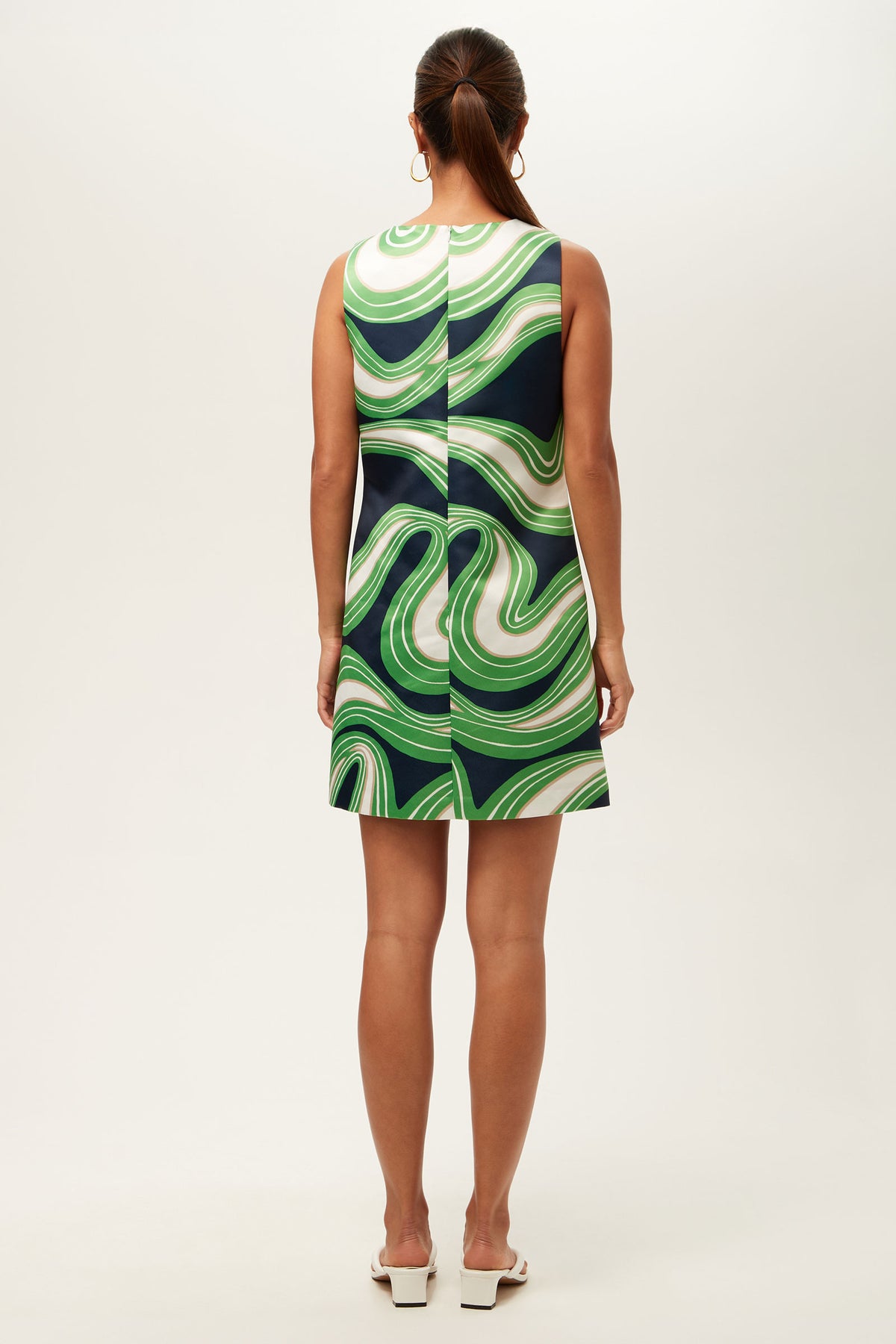 A woman with a low ponytail faces away, wearing the Fairy Tale 2 Sleeveless Dress - Serena Satin, a knee-length dress with bold green, white, and dark blue wavy patterns, paired with white heeled sandals.