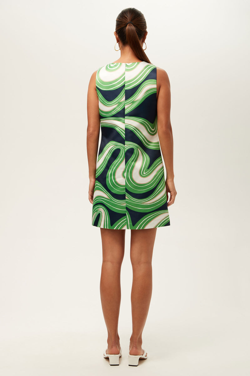 A woman with a low ponytail faces away, wearing the Fairy Tale 2 Sleeveless Dress - Serena Satin, a knee-length dress with bold green, white, and dark blue wavy patterns, paired with white heeled sandals.