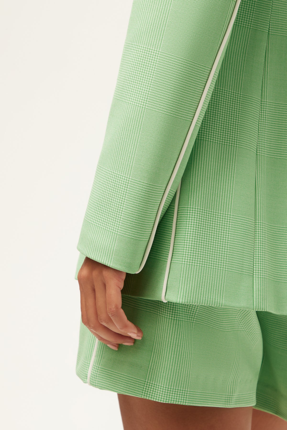 A close-up of a person wearing the Alternates Blazer - Game Time Glen Plaid, a textured light green blazer with white piping, and matching shorts, with their hand resting by their side against a plain background.