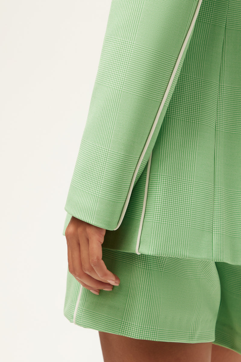 A close-up of a person wearing the Alternates Blazer - Game Time Glen Plaid, a textured light green blazer with white piping, and matching shorts, with their hand resting by their side against a plain background.
