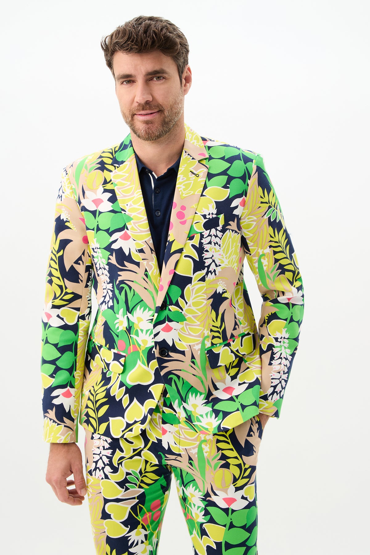 A man with short brown hair and a beard wears the Thurston Blazer - PS Eternal, featuring a bold, tropical floral pattern in green, yellow, pink, and navy. He stands against a plain white background, looking at the camera with a slight smile.