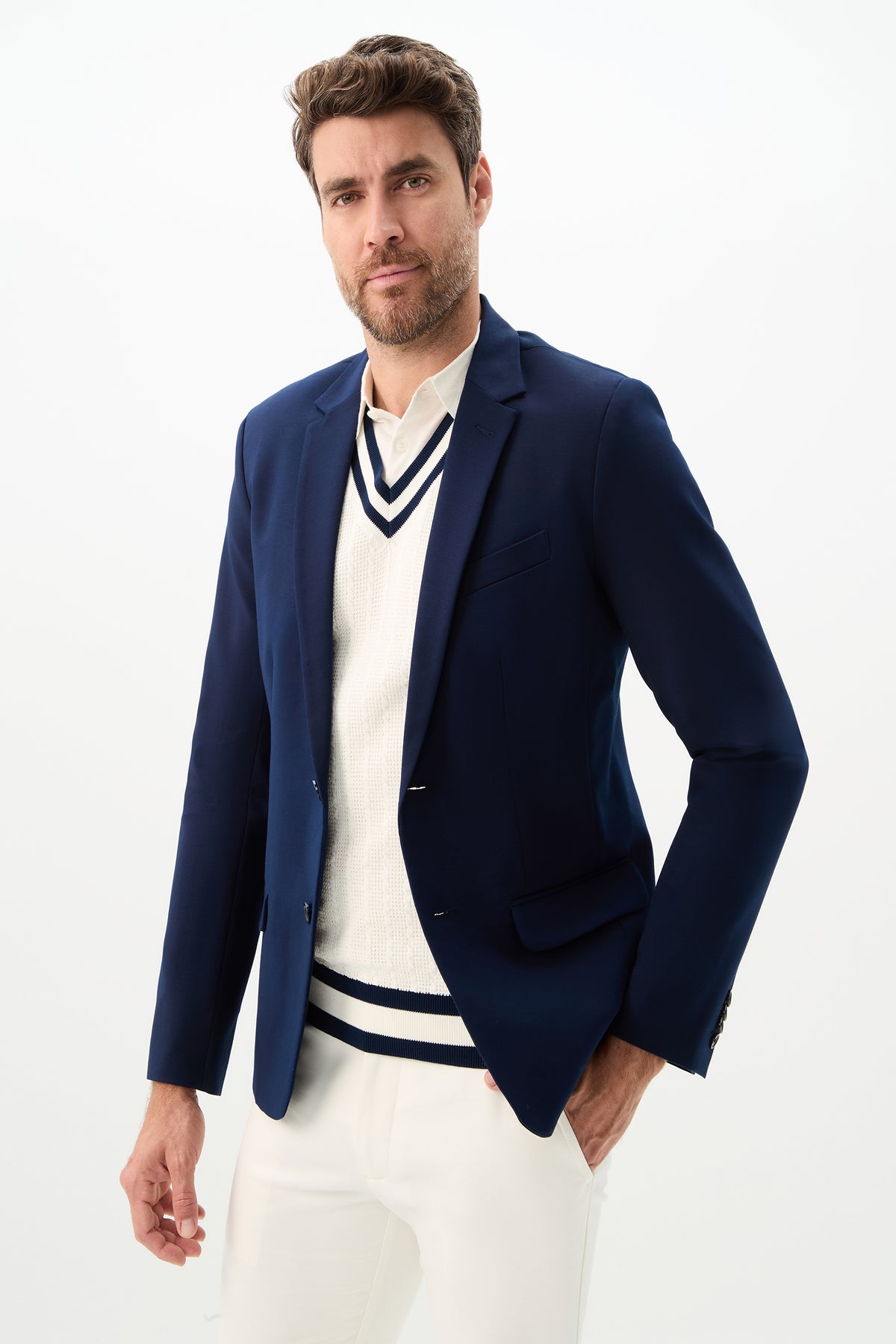 A man with short brown hair and a beard confidently poses in the Thurston Blazer - Solid Suiting, styled over a white sweater with navy trim and white pants, against a plain white background.