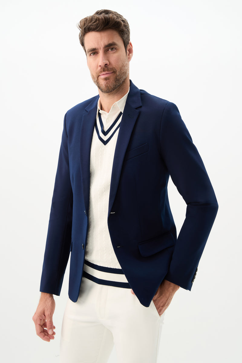 A man with short brown hair and a beard confidently poses in the Thurston Blazer - Solid Suiting, styled over a white sweater with navy trim and white pants, against a plain white background.