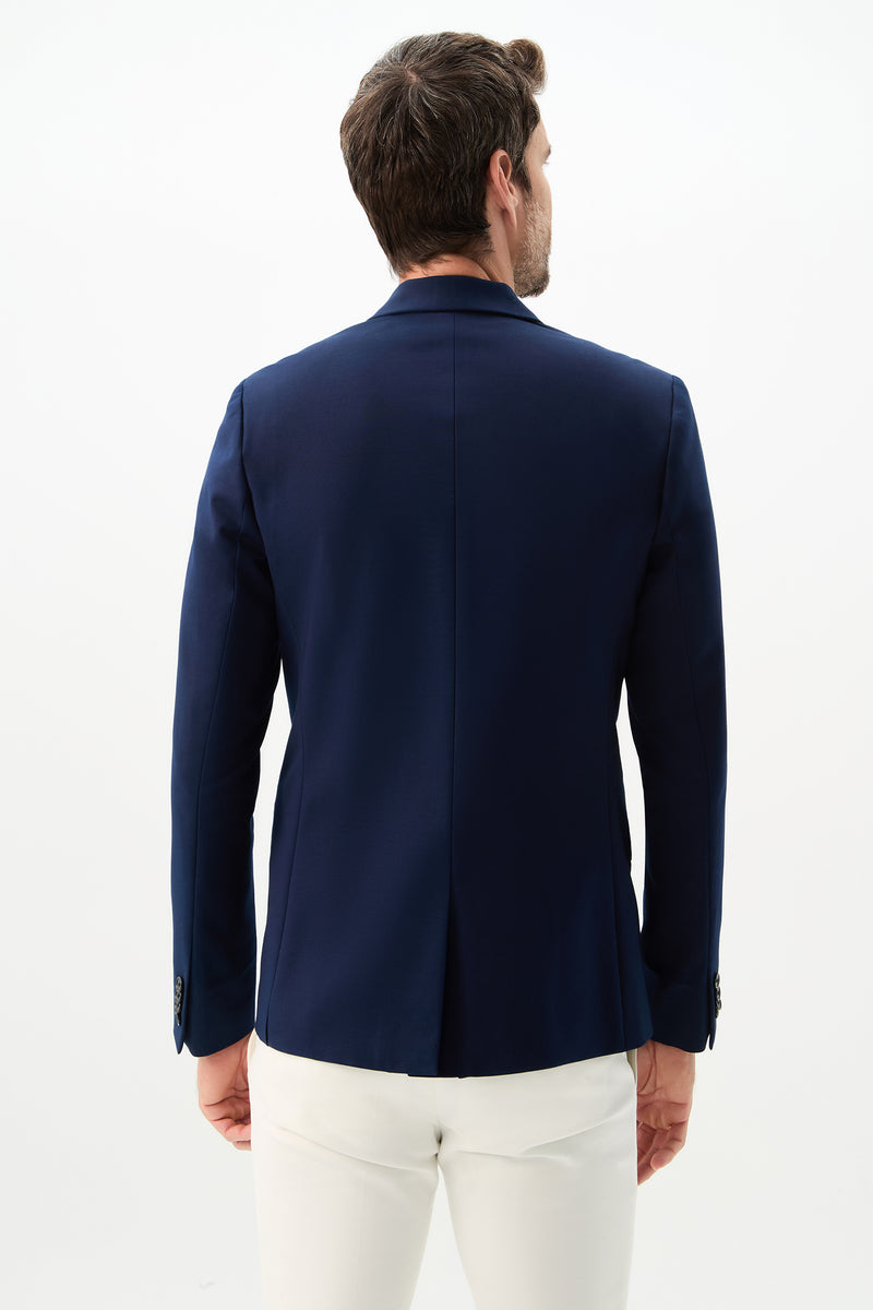 A man is seen from behind wearing the Thurston Blazer - Solid Suiting in navy blue with white pants, standing against a plain white background.