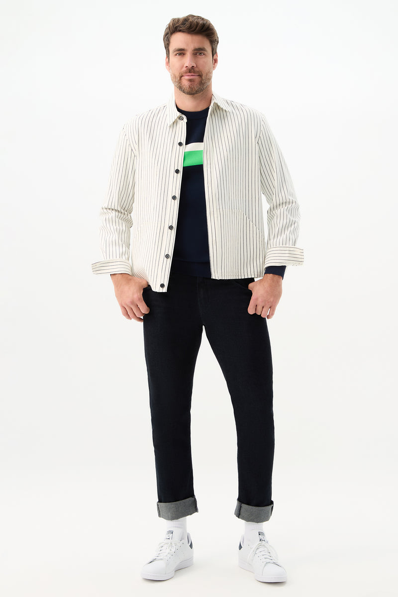 A man faces forward against a plain white background, wearing the Mulholland Shirt Jacket in Seles Stripe over a dark shirt with a green stripe, paired with cuffed dark jeans and white sneakers.