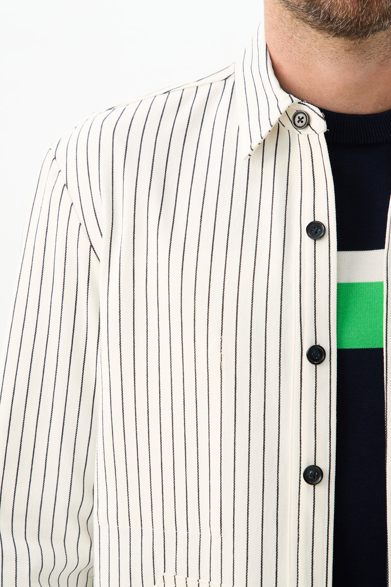 A close-up of a person wearing the Mulholland Shirt Jacket - Seles Stripe, a white shirt with black vertical stripes, over a dark sweater with white and green horizontal stripes. The shirt is unbuttoned at the top, partially revealing a bearded face.