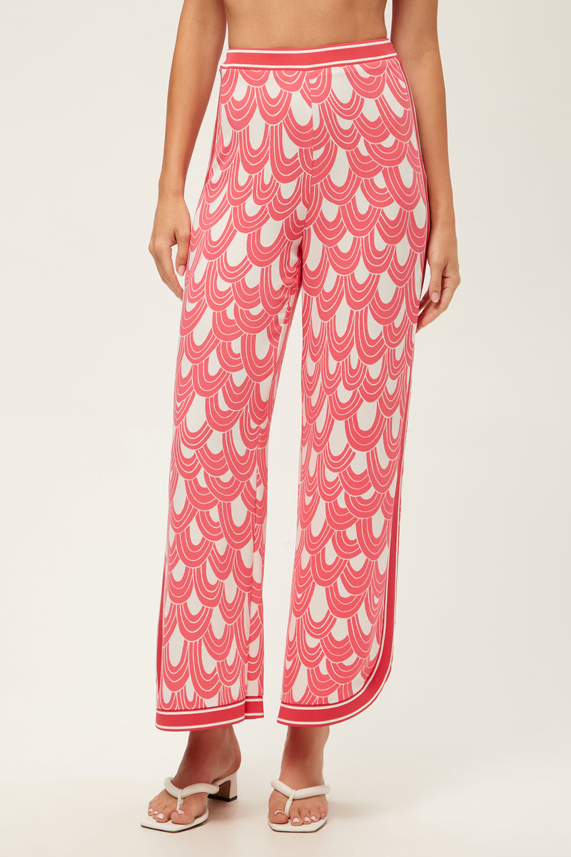 A woman showcases the Overhead Wide-Leg Pant - Ace Arches in Coral Crush/Whitewash, featuring a scalloped pattern and high-waisted, wide-leg fit. She pairs them with white heeled sandals; the image is cropped from shoulders down on a plain background.