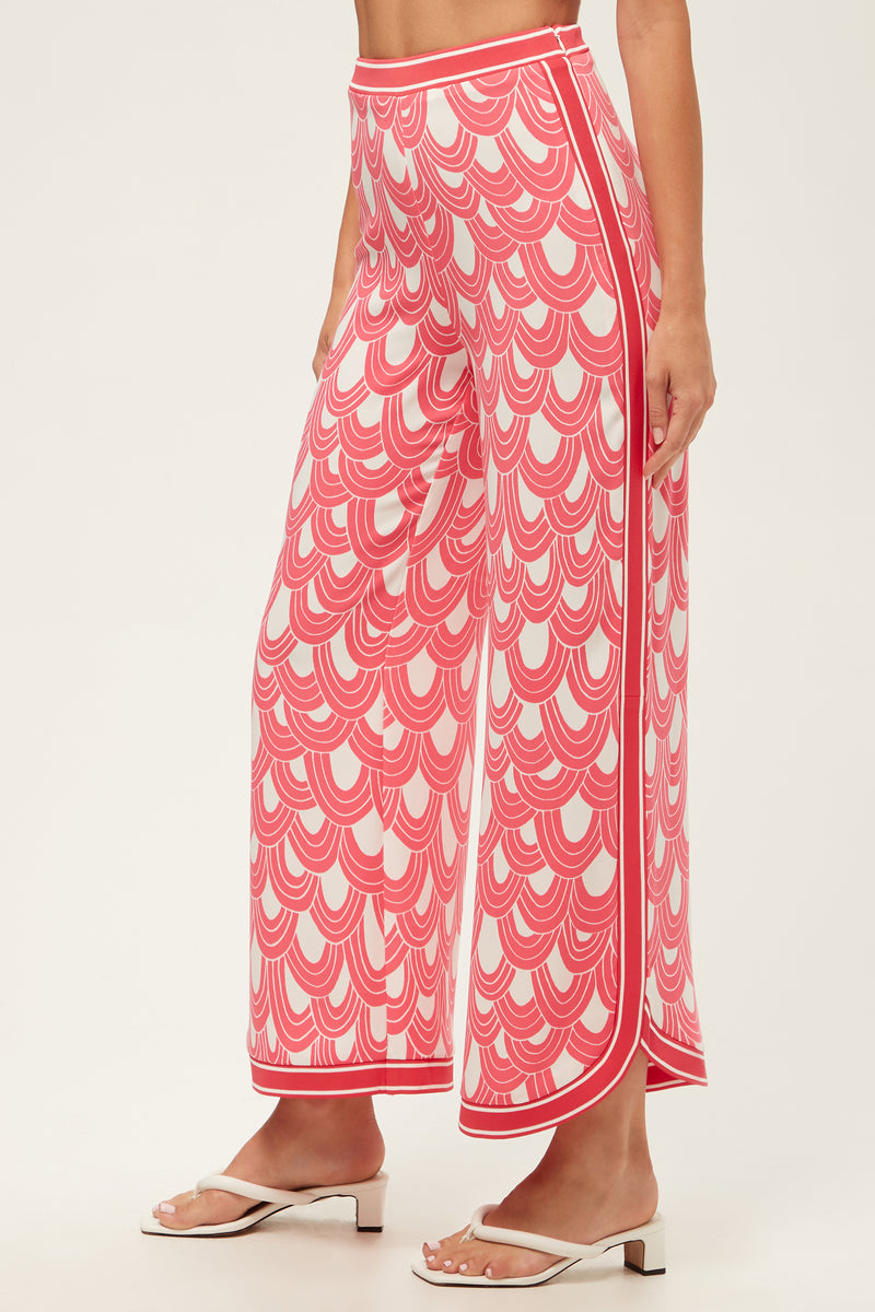 A person wears the Overhead Wide-Leg Pant - Ace Arches, which features a pink and white scalloped pattern, side stripes, and a split hem detail, paired with white heeled sandals.
