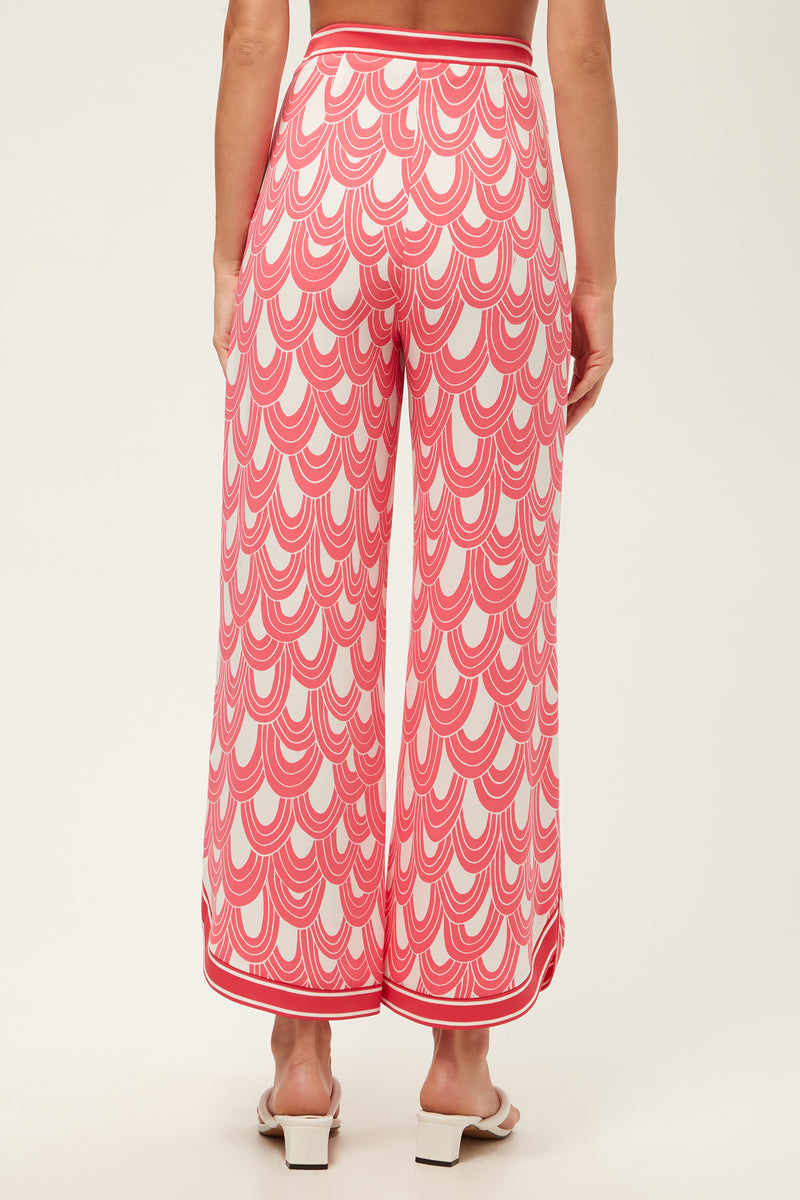 A person wears the Overhead Wide-Leg Pant - Ace Arches, featuring a pink and white scallop pattern, standing barefoot and facing away from the camera against a plain background.