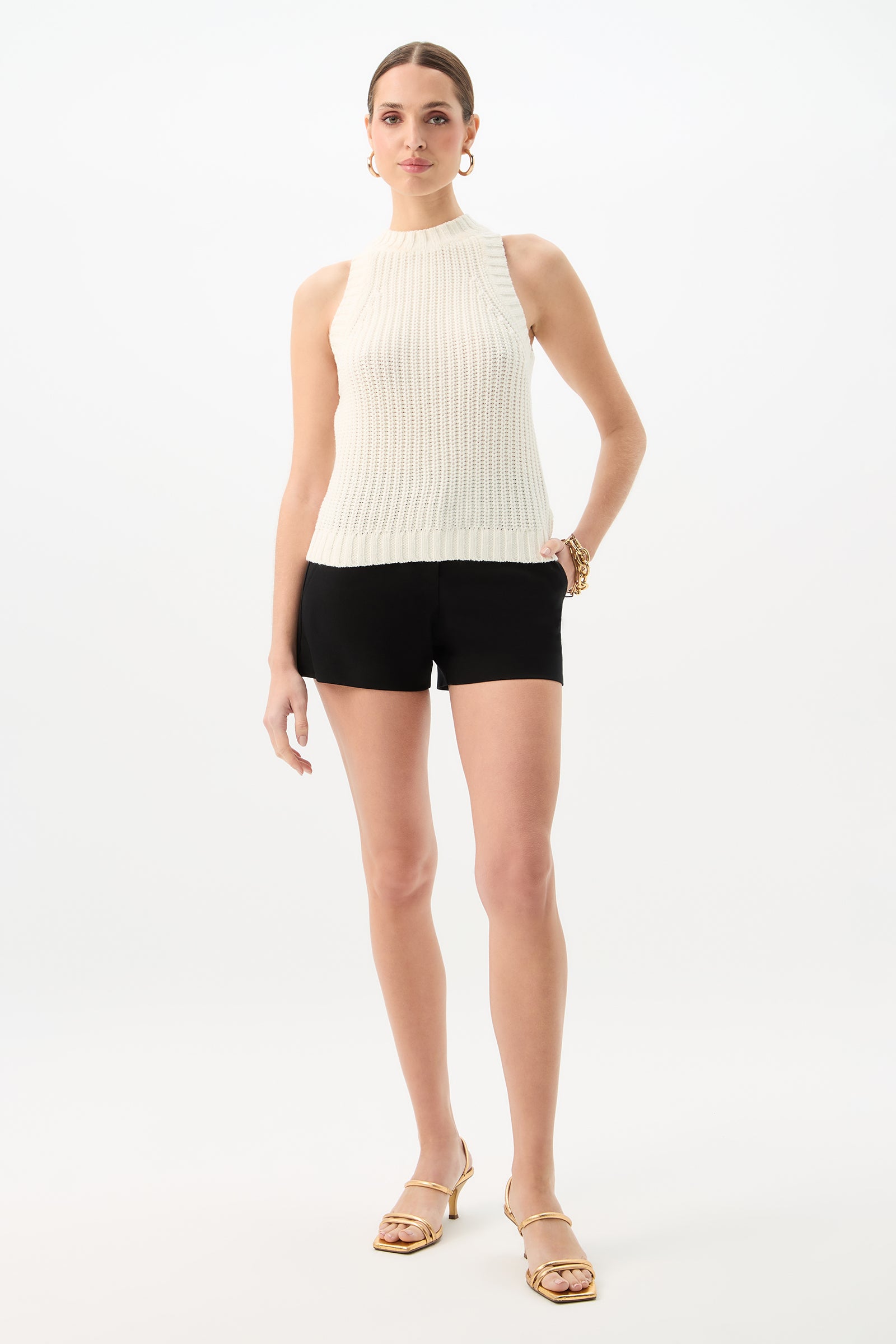 A woman poses against a white background wearing Corbin 2 Shorts - Cotton Bi-Stretch in black, paired with a sleeveless white ribbed top, gold hoop earrings, bracelets, and gold heeled sandals. She has her hair pulled back and one hand on her hip.