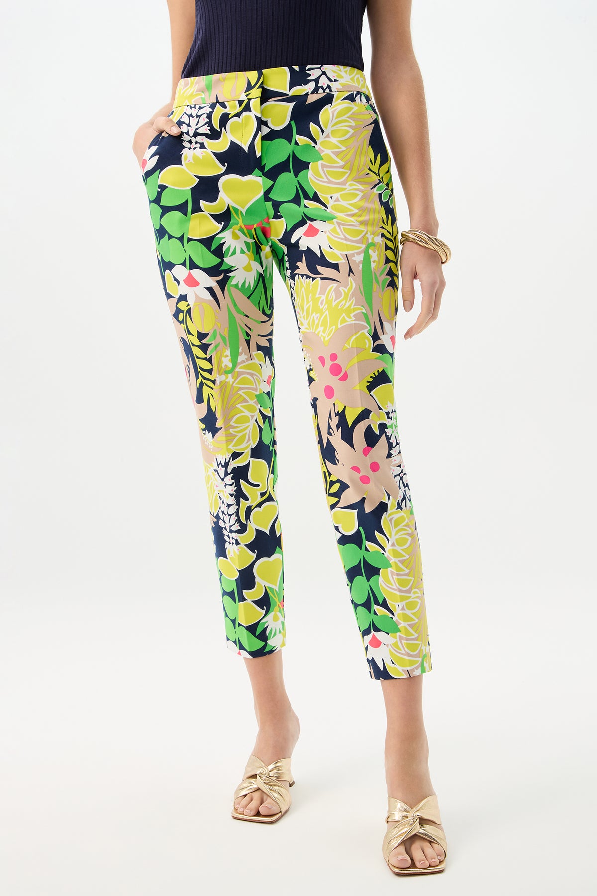 A woman wears the Aubree 2 Pant - PS Eternal in Palm Springs Eternal, featuring vibrant florals, paired with a sleeveless black top, gold sandals, and gold bangles, with one hand in her pocket.