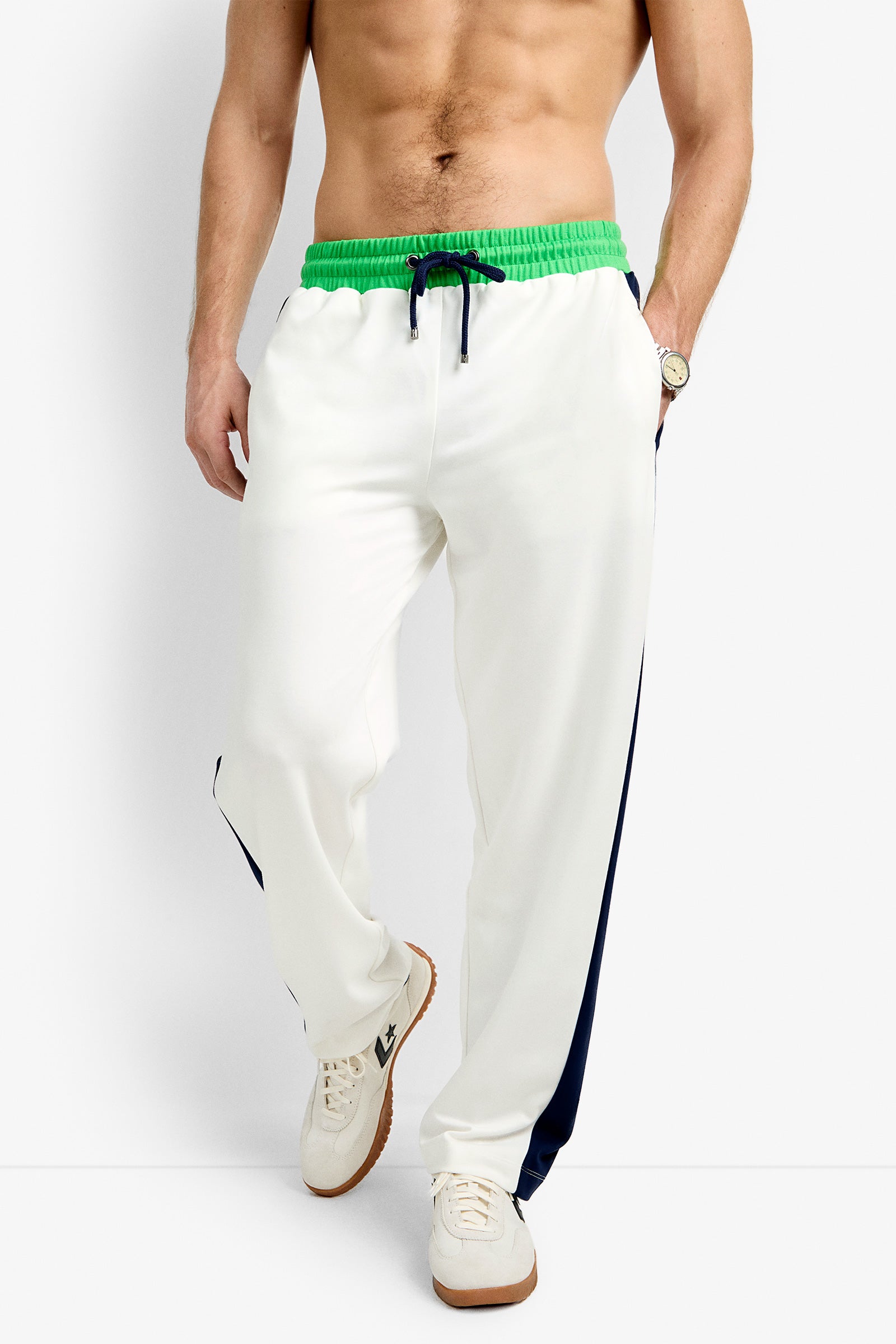 A man wearing the Tennis Track Pant - Cozy Ponte, featuring a green drawstring waistband and navy side stripes, paired with white sneakers and a wristwatch, stands against a plain background.