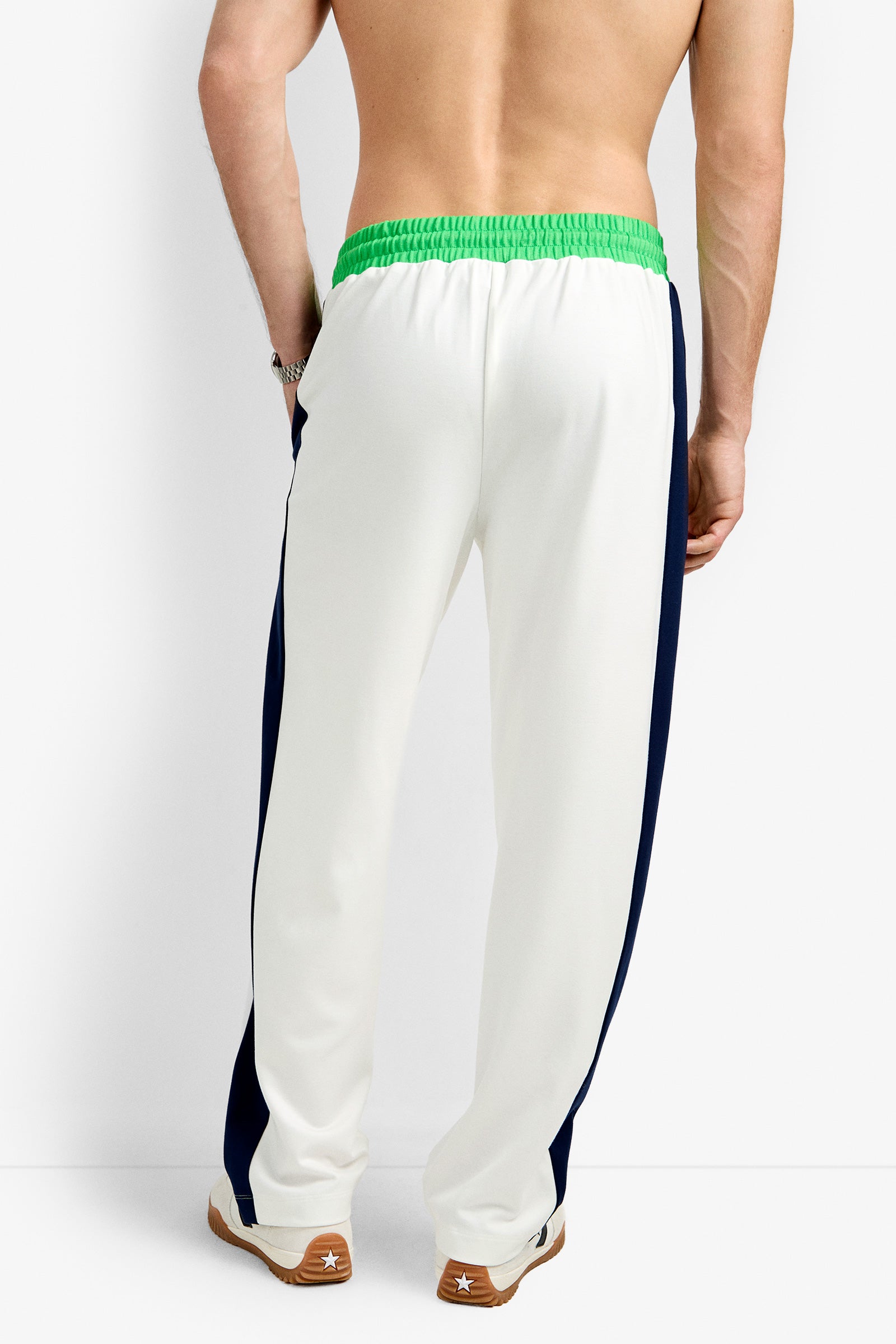 A shirtless man faces away, wearing the Tennis Track Pant - Cozy Ponte in white with a green elastic waistband, navy side stripes, and brown sneakers with white soles, set against a plain white background.